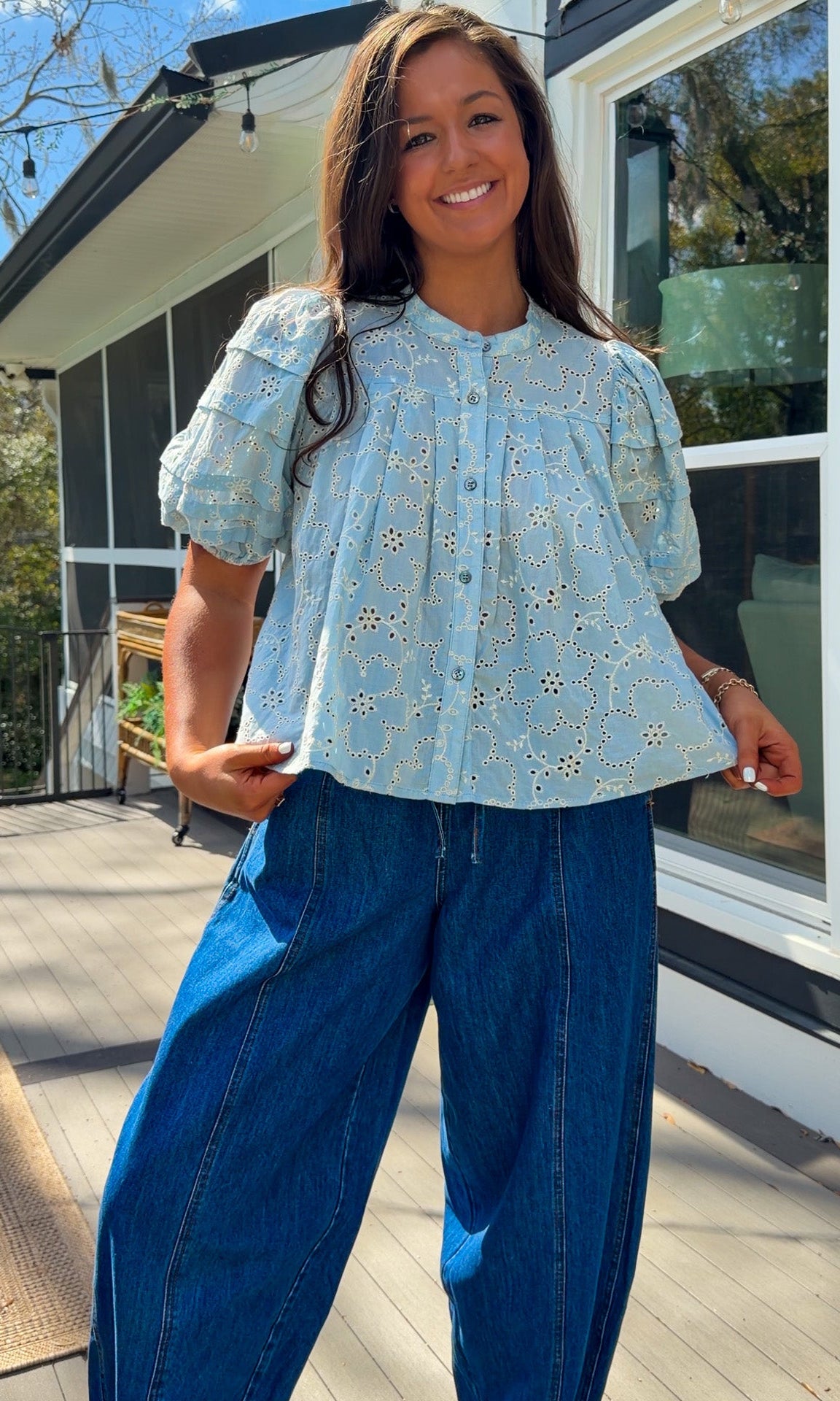 Woman wearing a yellow blouse with a pattern and blue jeans standing on a wooden deck.