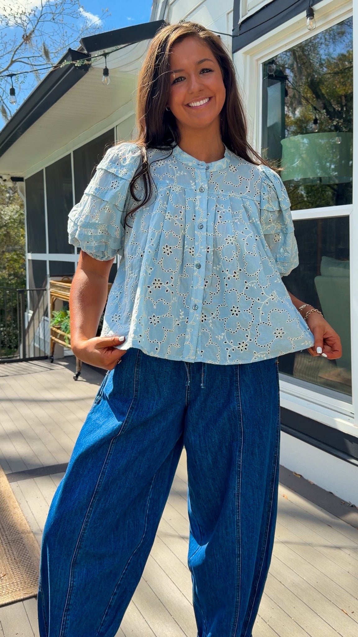 Woman wearing a yellow blouse with a pattern and blue jeans standing on a wooden deck.