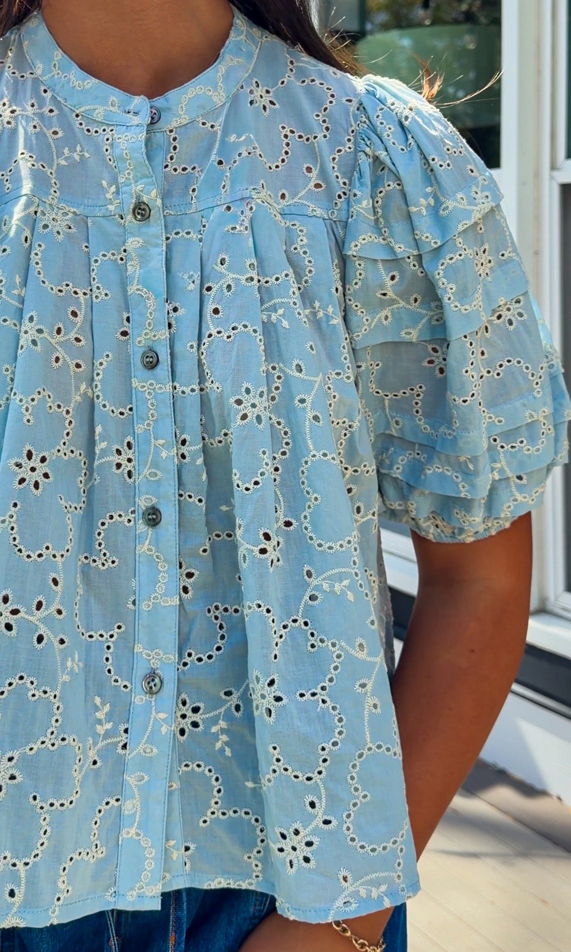 Woman wearing a yellow blouse with a pattern and blue jeans standing on a wooden deck.