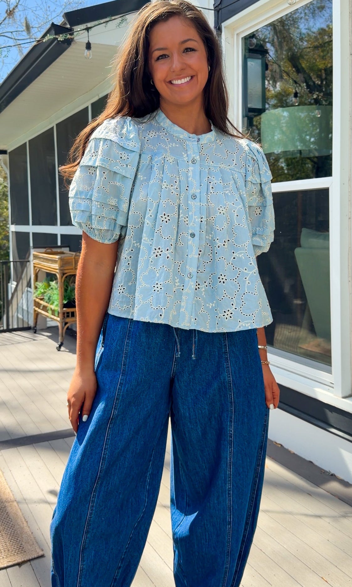 Woman wearing a light blue blouse with floral patterns and blue jeans standing outdoors.
