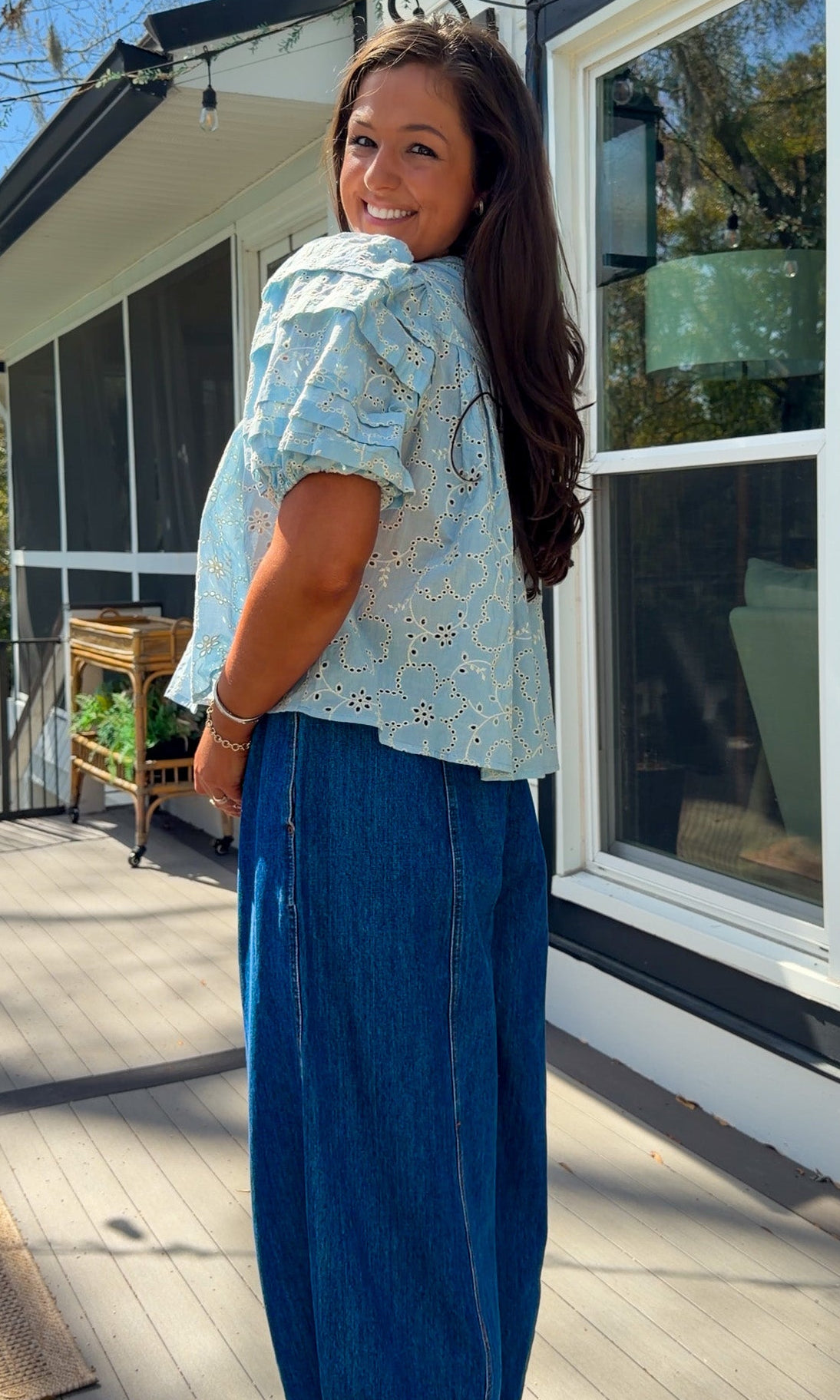 Woman wearing a light blue blouse with floral patterns and blue jeans standing outdoors.