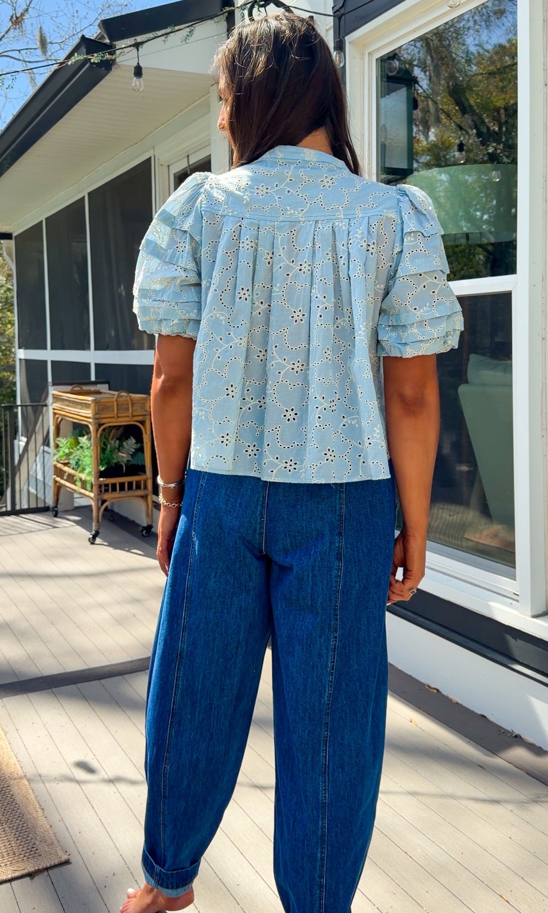 Woman wearing a light blue blouse with floral patterns and blue jeans standing outdoors.