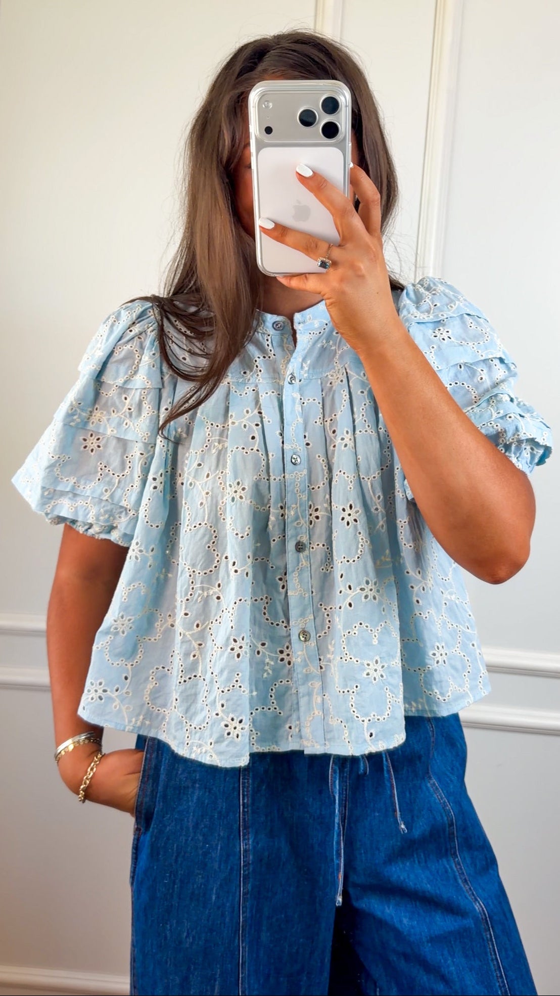 Woman wearing a light blue blouse with floral patterns and blue jeans standing outdoors.