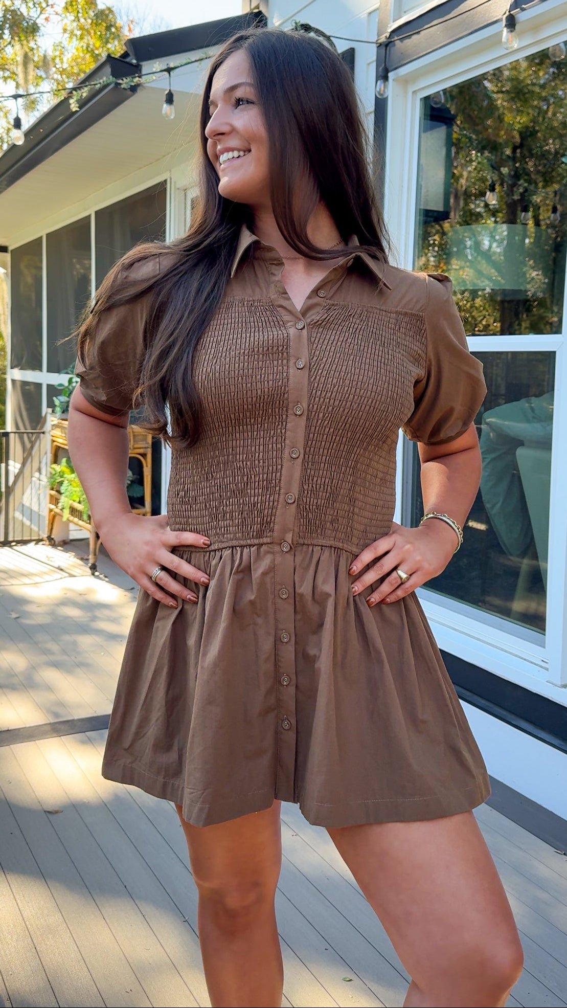 Woman in a brown dress standing on a wooden deck with a house in the background
