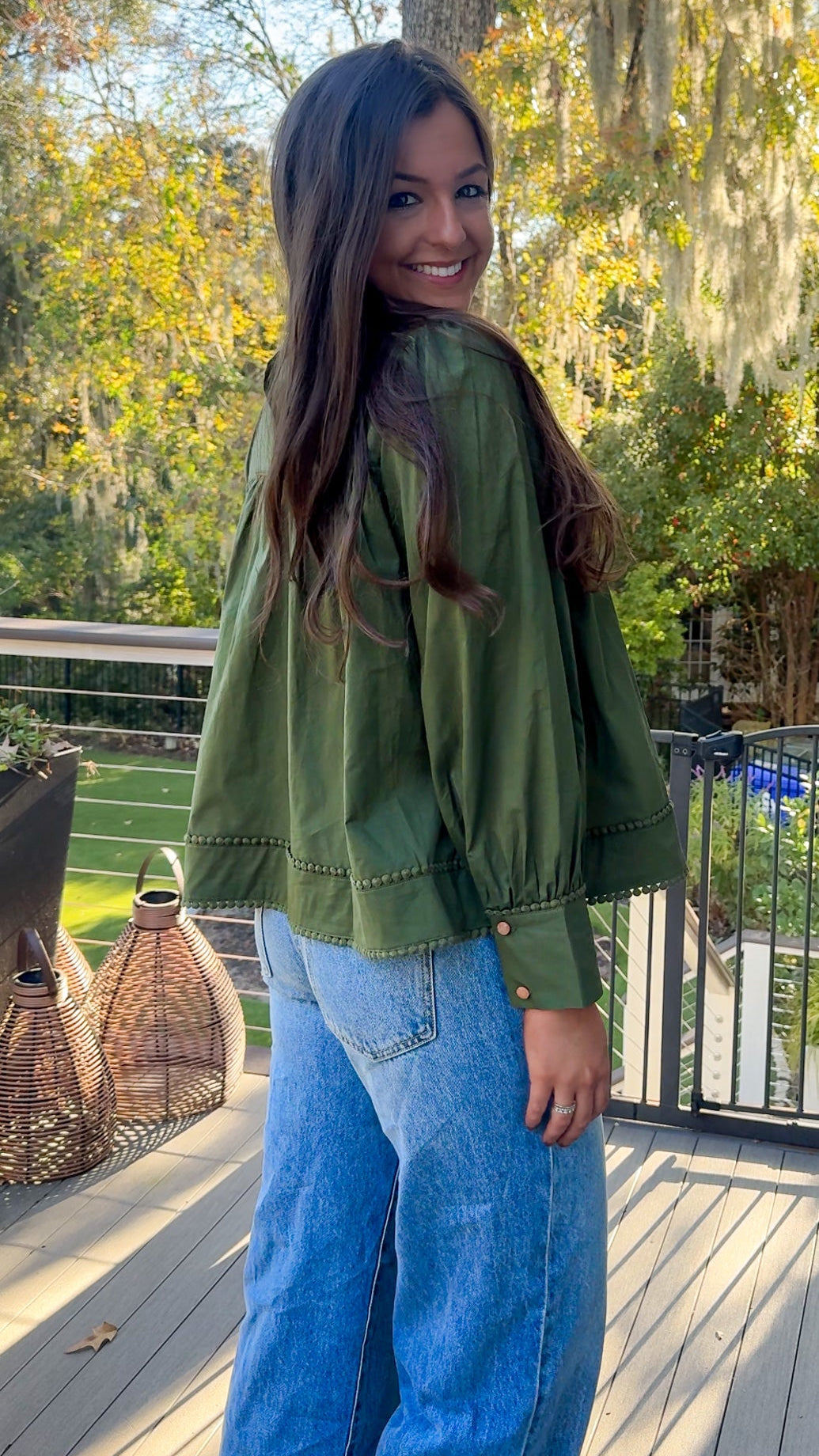 Woman wearing a green jacket and blue jeans standing on a wooden deck with trees in the background