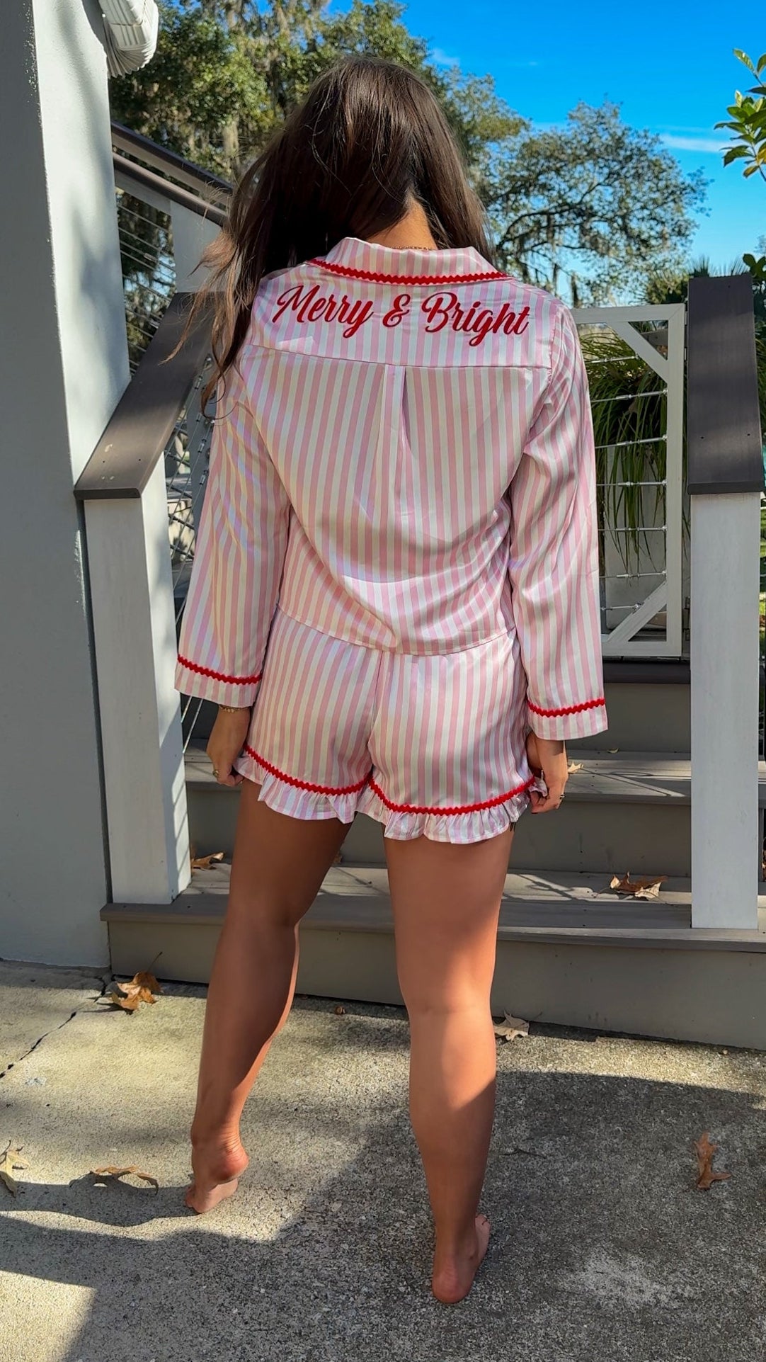 Woman wearing a pink and white striped pajama set with red accents outdoors. christmas pajamas merry and bright wording on back