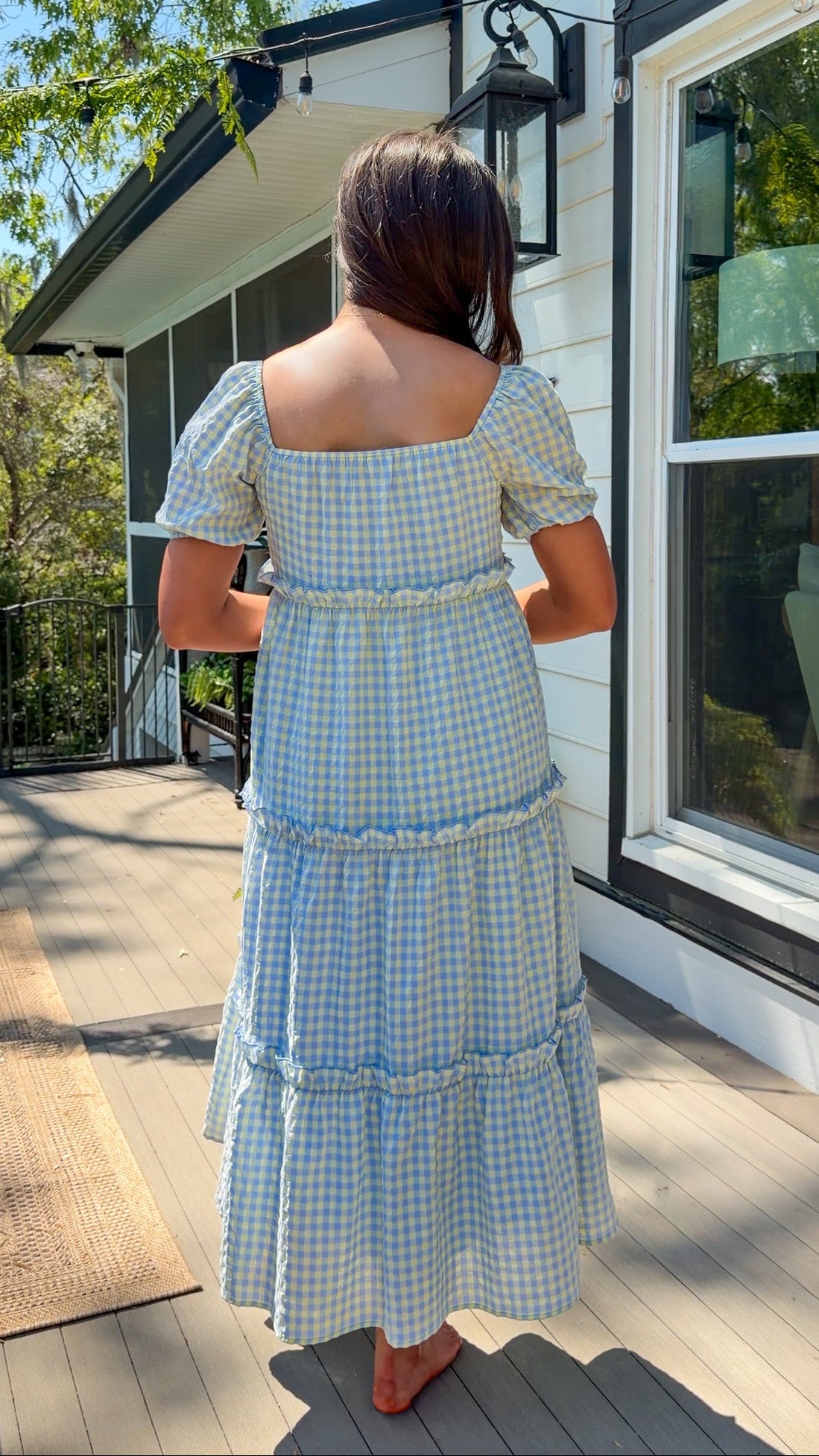 Woman in a blue checkered dress standing on a wooden deck with a house in the background.