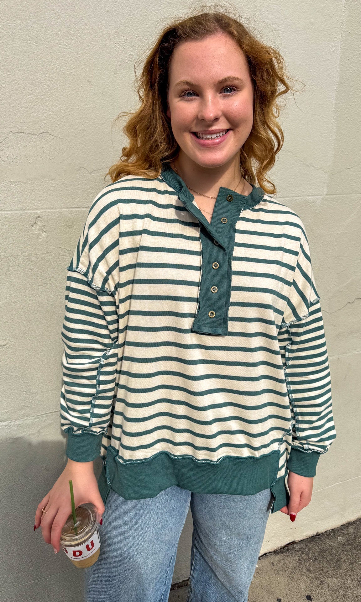 Woman wearing a striped shirt and jeans, holding a drink against a plain wall.