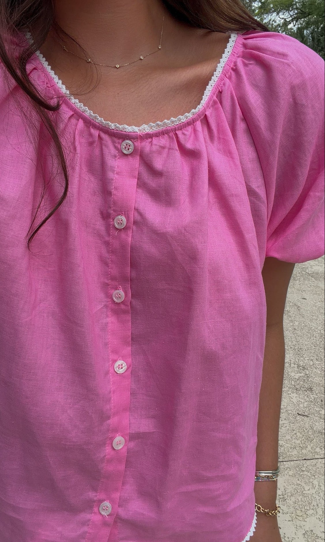 A close-up of a woman wearing a pink short-sleeve button-front blouse with white lace trim around the neckline and hem.