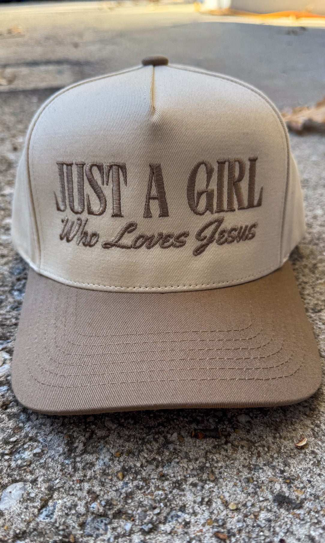 Beige cap with 'Just a Girl Who Loves Jesus' text on a concrete surface