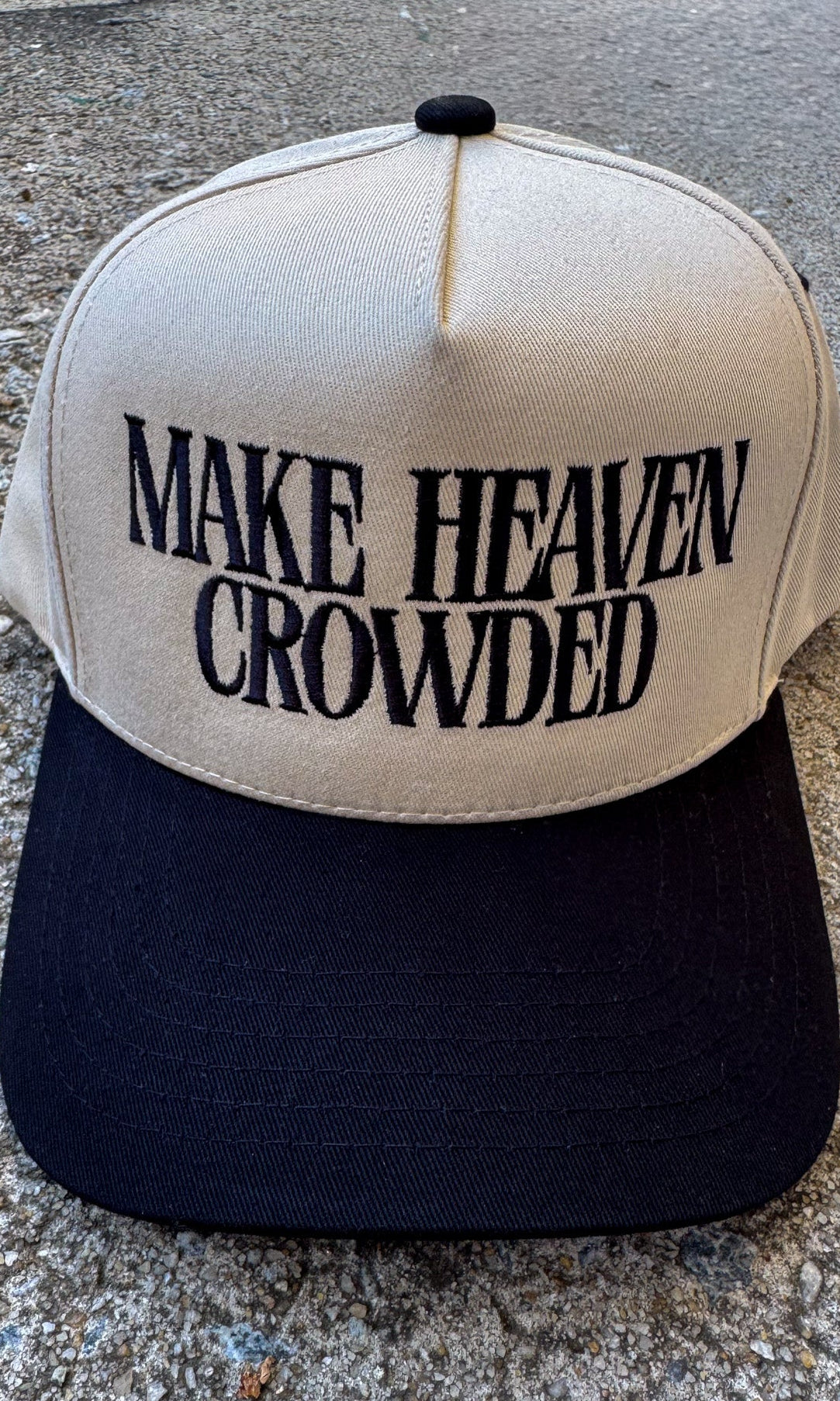 Beige and black cap with 'MAKE HEAVEN CROWDED' text on a concrete surface