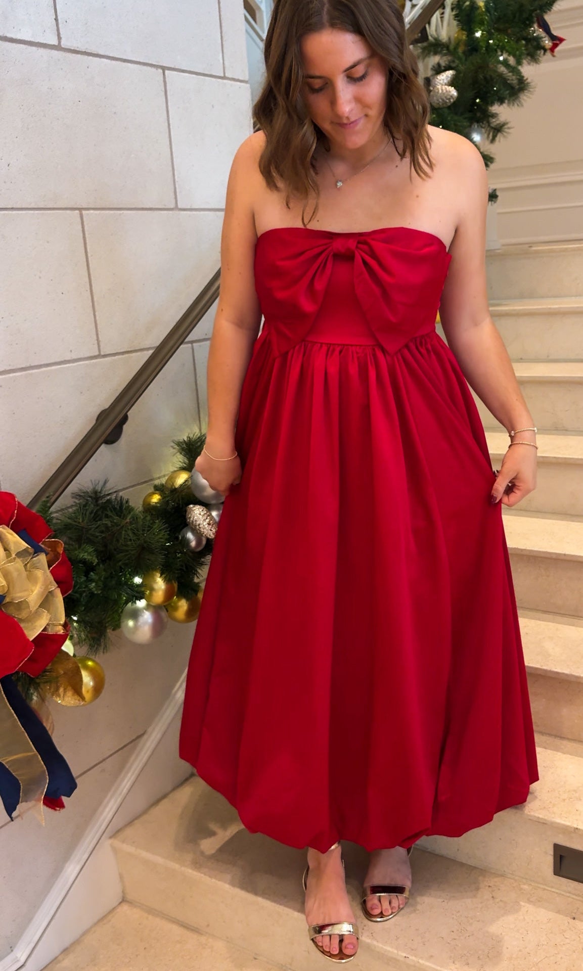 Red strapless midi dress with large front bow detail, full gathered skirt, and smocked back panel, worn on staircase with holiday decor.