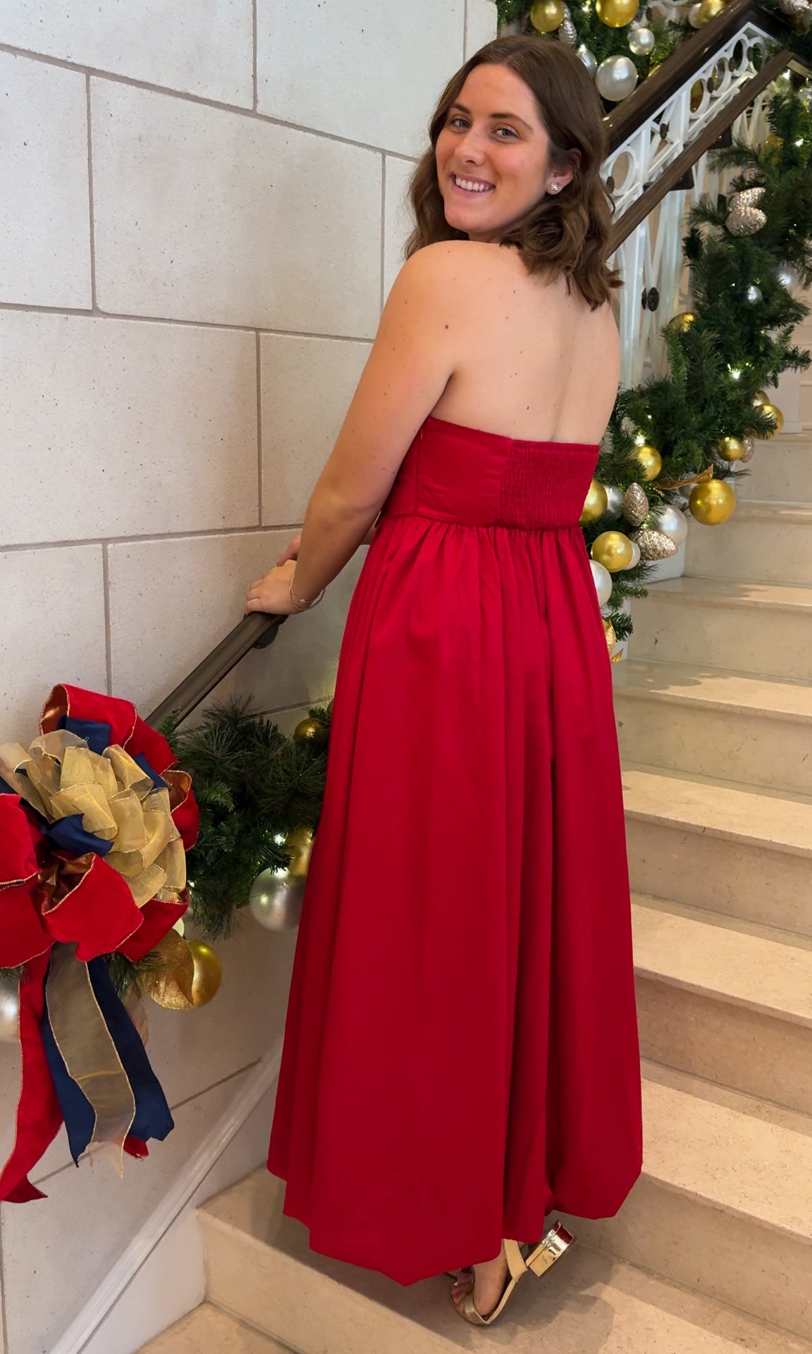 Red strapless midi dress with large front bow detail, full gathered skirt, and smocked back panel, worn on staircase with holiday decor.