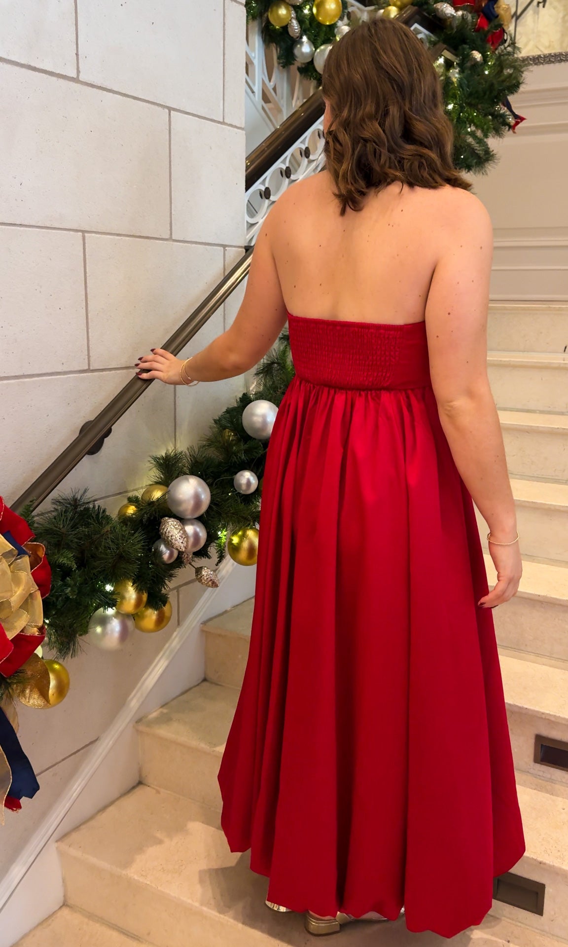 Red strapless midi dress with large front bow detail, full gathered skirt, and smocked back panel, worn on staircase with holiday decor.