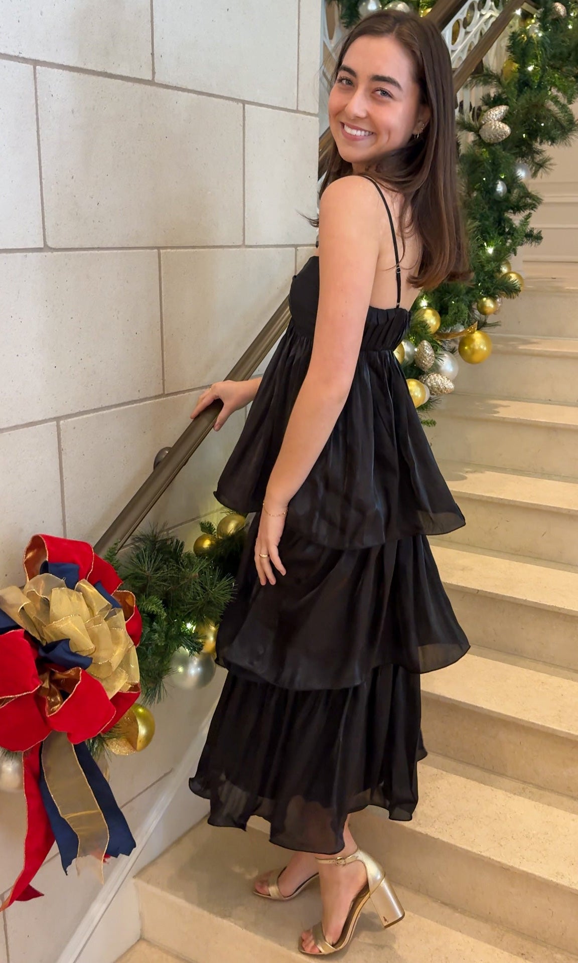 Black tiered midi dress with spaghetti straps, sweetheart-style neckline, and back zipper closure, worn on indoor staircase with holiday decor.