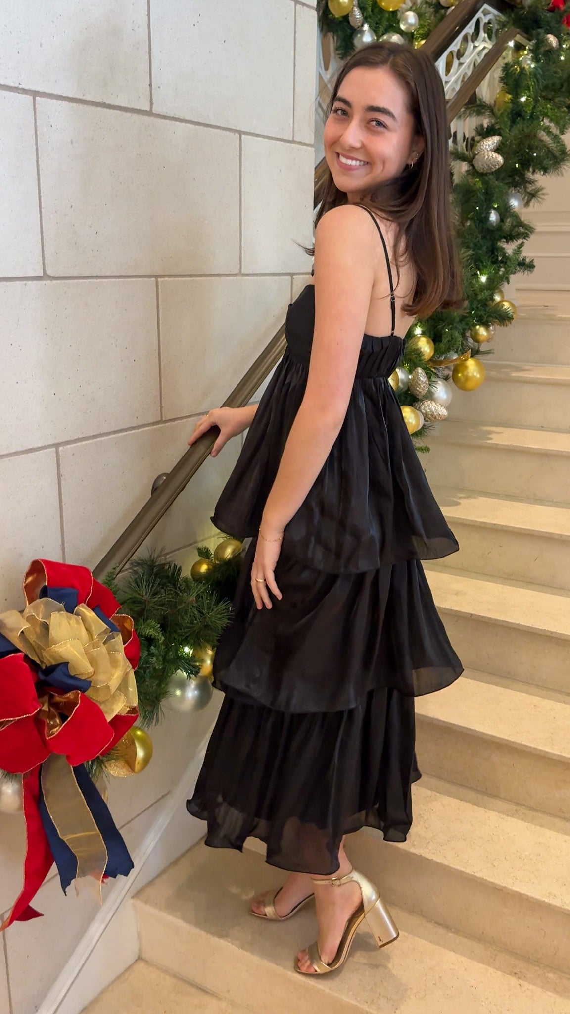 Black tiered midi dress with spaghetti straps, sweetheart-style neckline, and back zipper closure, worn on indoor staircase with holiday decor.