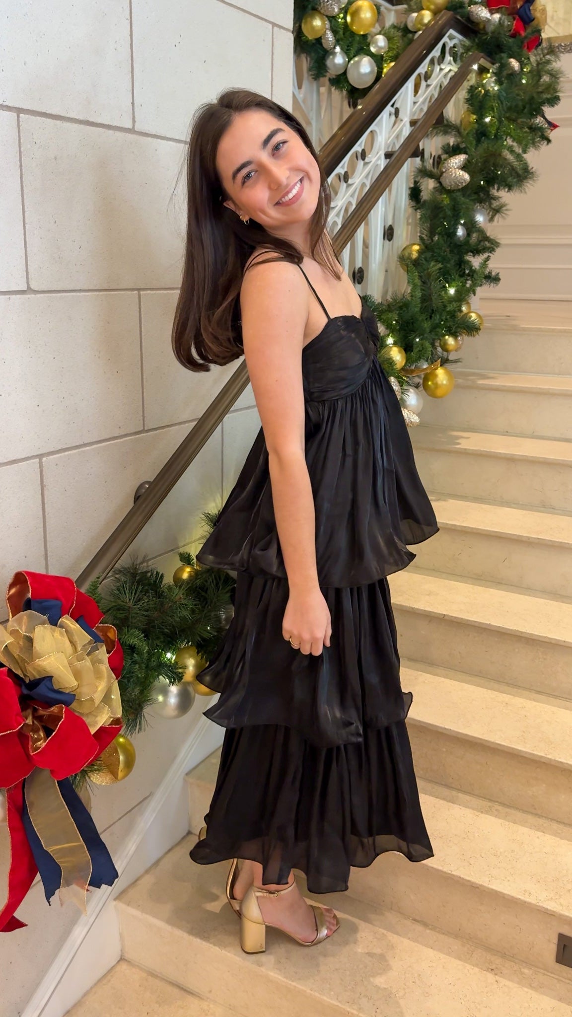 Black tiered midi dress with spaghetti straps, sweetheart-style neckline, and back zipper closure, worn on indoor staircase with holiday decor.