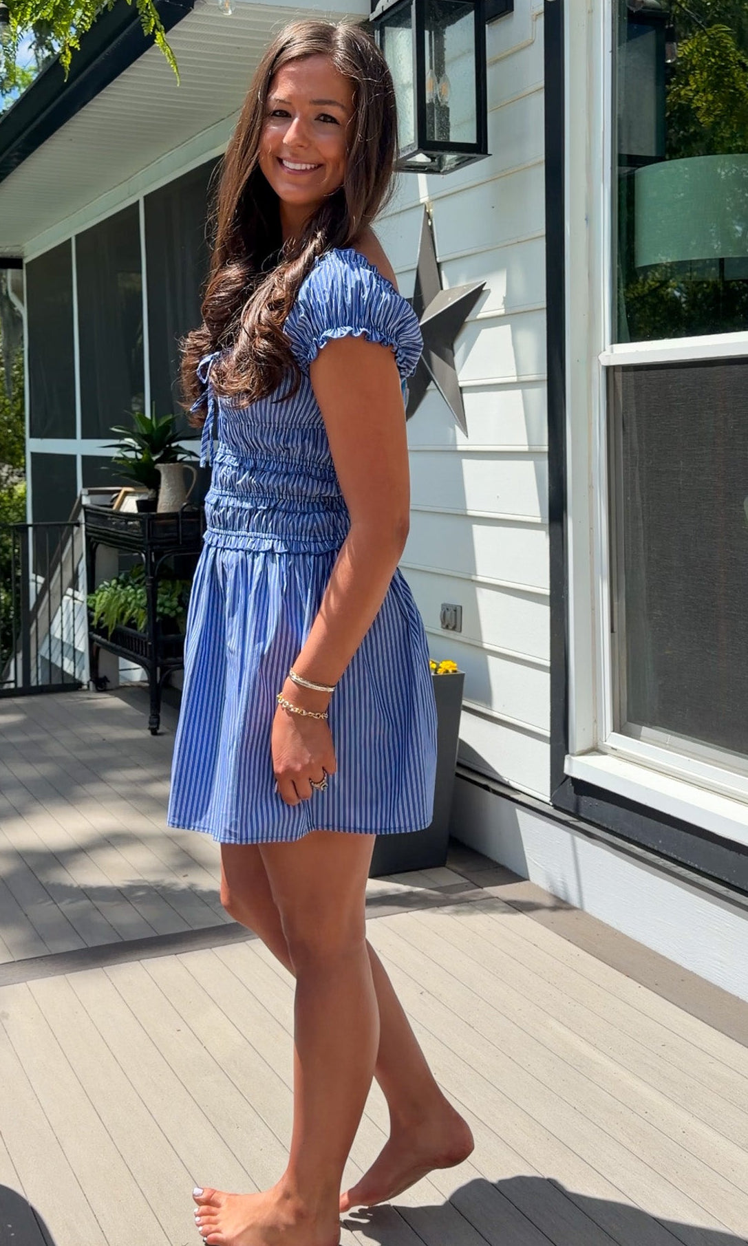 Woman in a blue dress standing on a wooden deck outside a house.