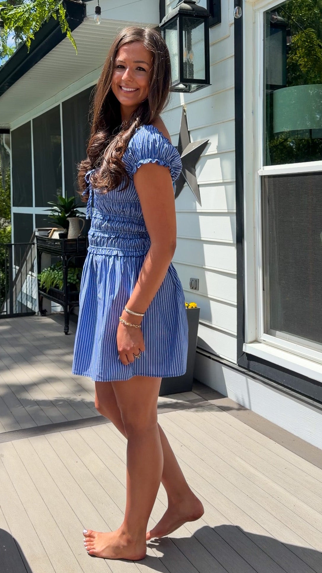 Woman in a blue dress standing on a wooden deck outside a house.
