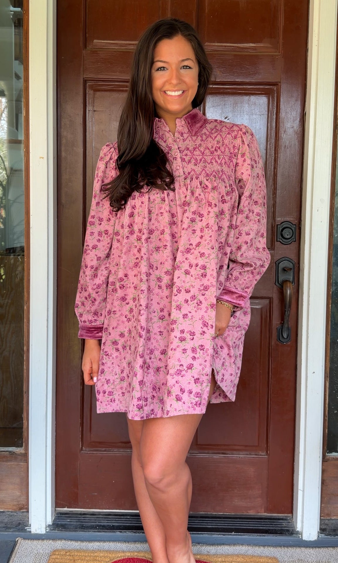 Model wearing a mauve floral corduroy babydoll dress with smocked bodice and velvet collar and cuffs standing in front of a wooden door.