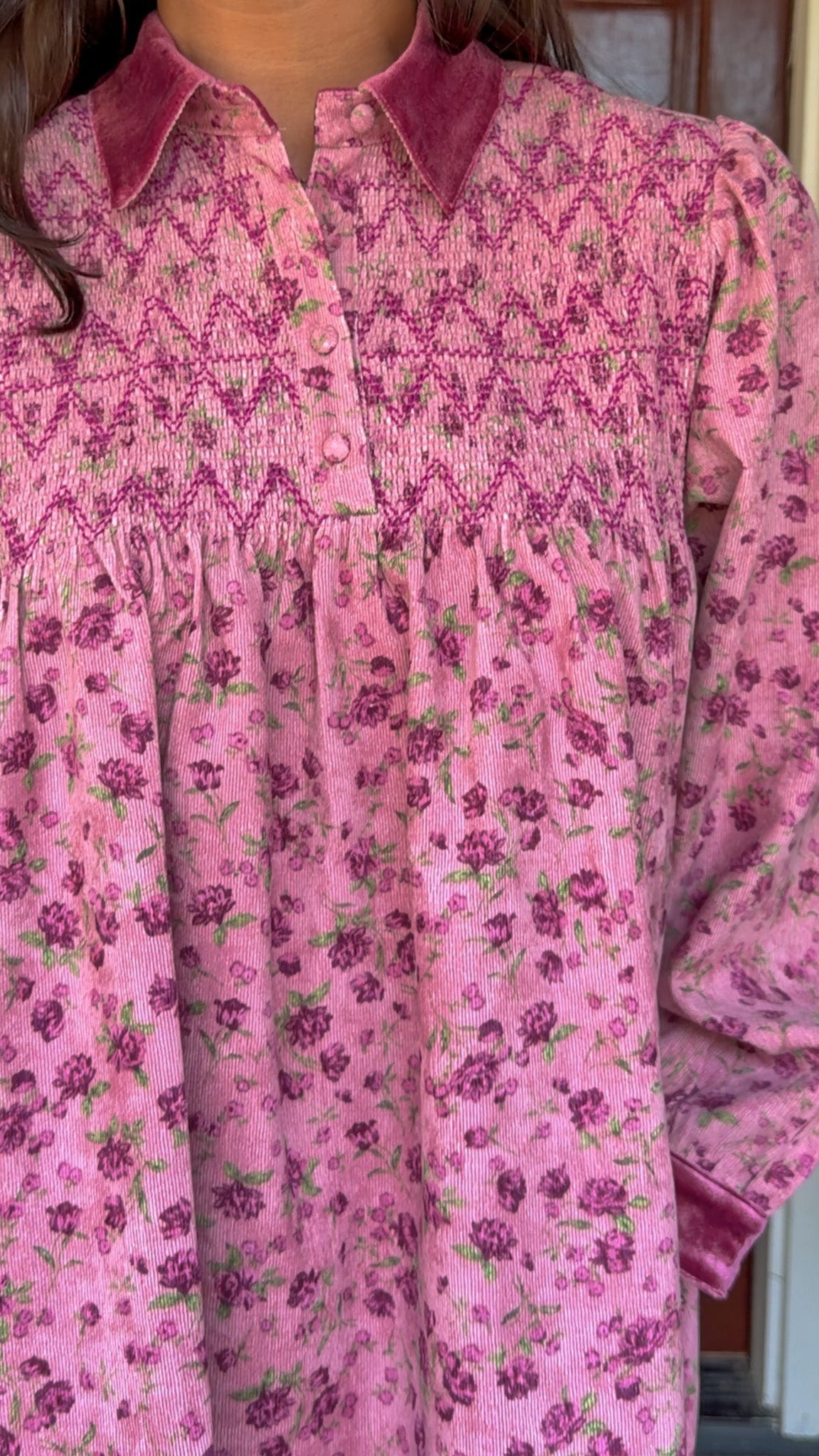Model wearing a mauve floral corduroy babydoll dress with smocked bodice and velvet collar and cuffs standing in front of a wooden door.