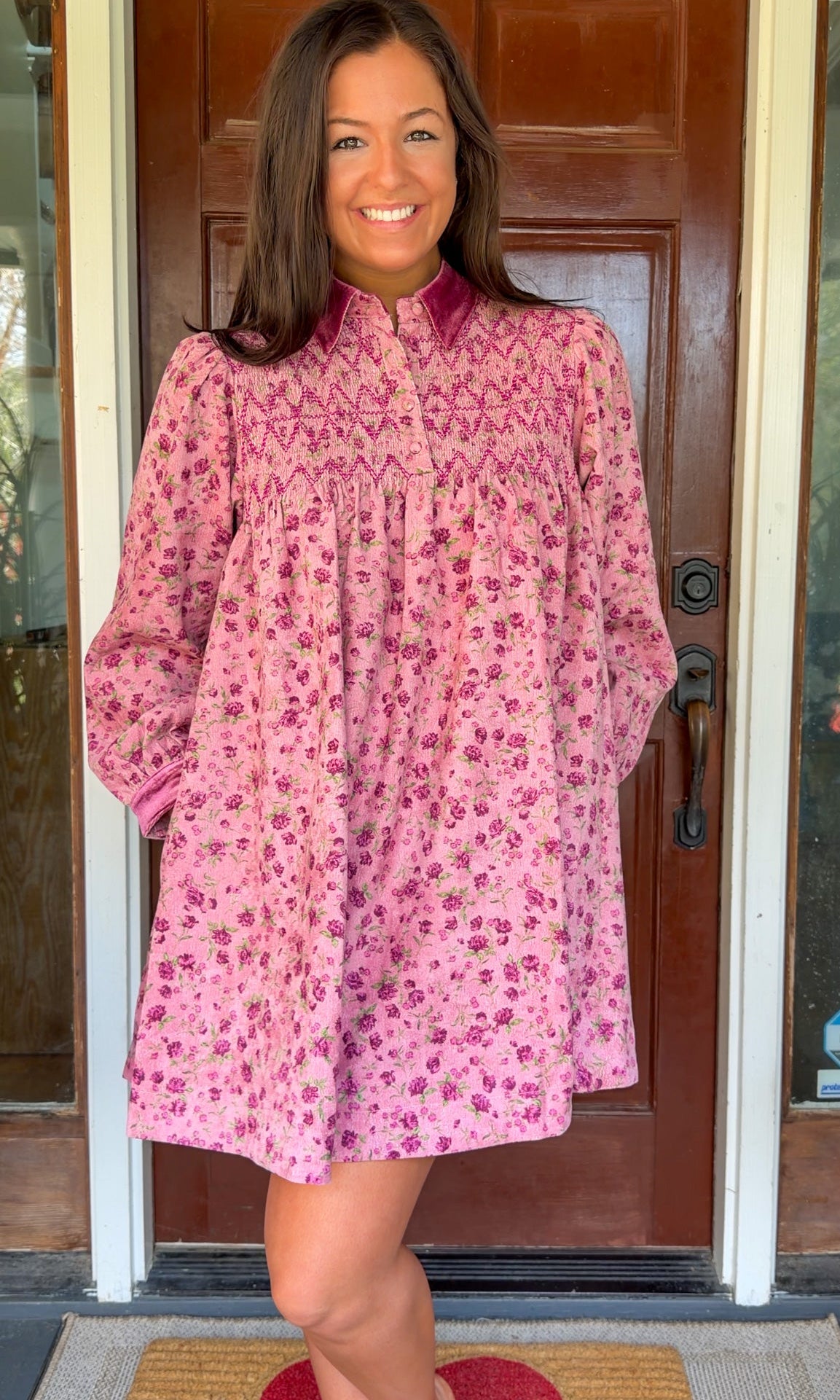 Model wearing a mauve floral corduroy babydoll dress with smocked bodice and velvet collar and cuffs standing in front of a wooden door.