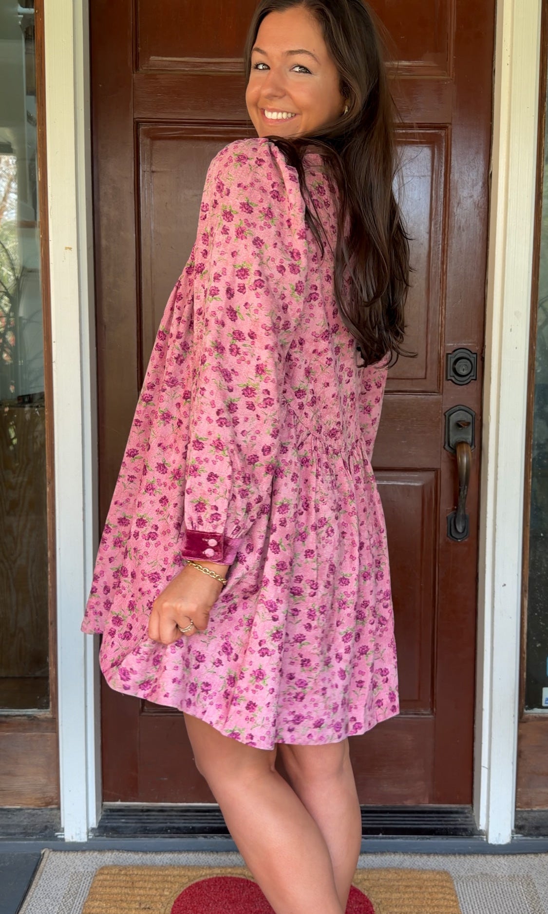 Model wearing a mauve floral corduroy babydoll dress with smocked bodice and velvet collar and cuffs standing in front of a wooden door.