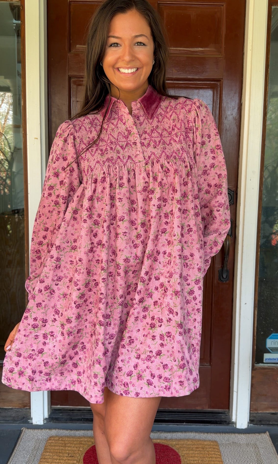 Model wearing a mauve floral corduroy babydoll dress with smocked bodice and velvet collar and cuffs standing in front of a wooden door.