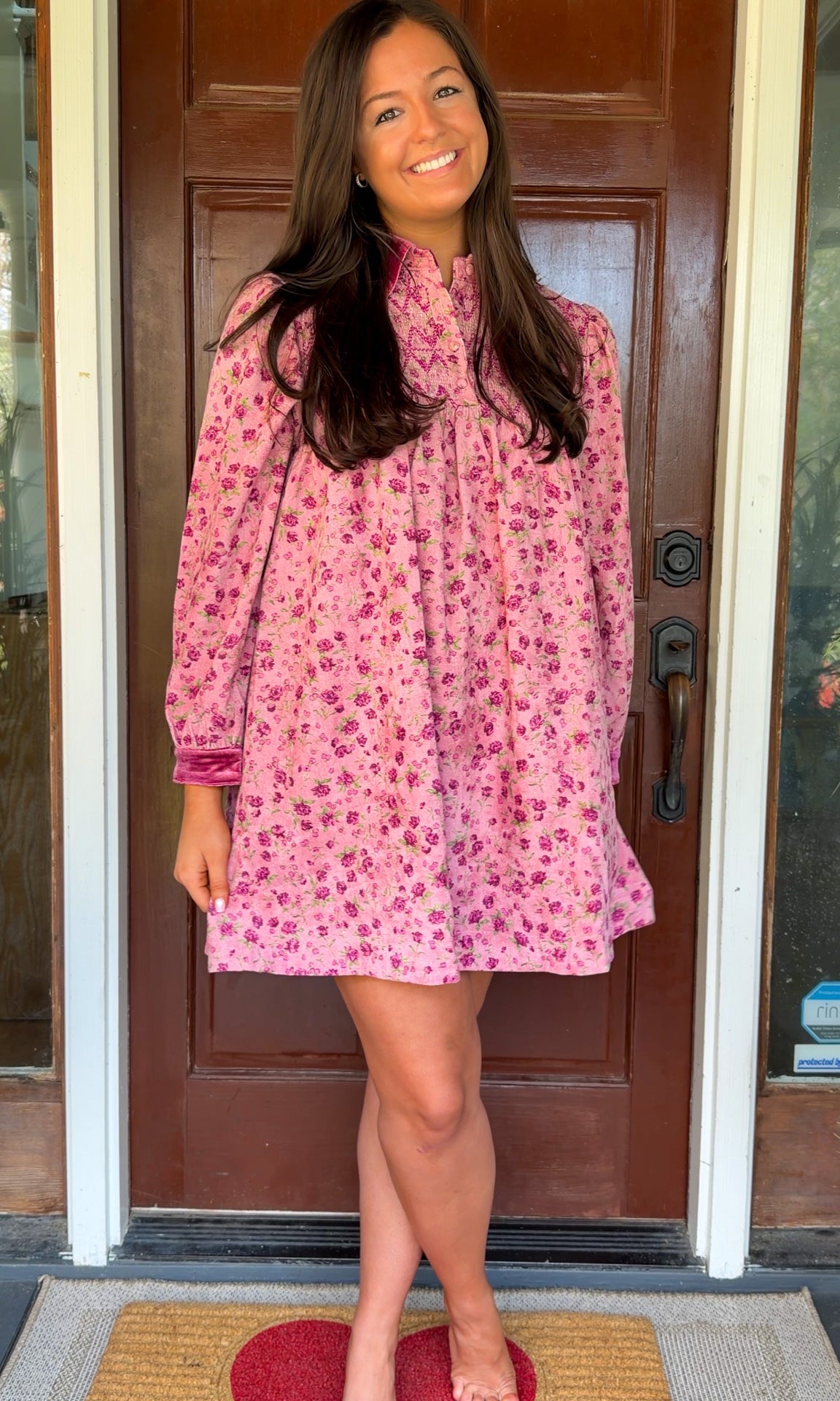 Model wearing a mauve floral corduroy babydoll dress with smocked bodice and velvet collar and cuffs standing in front of a wooden door.