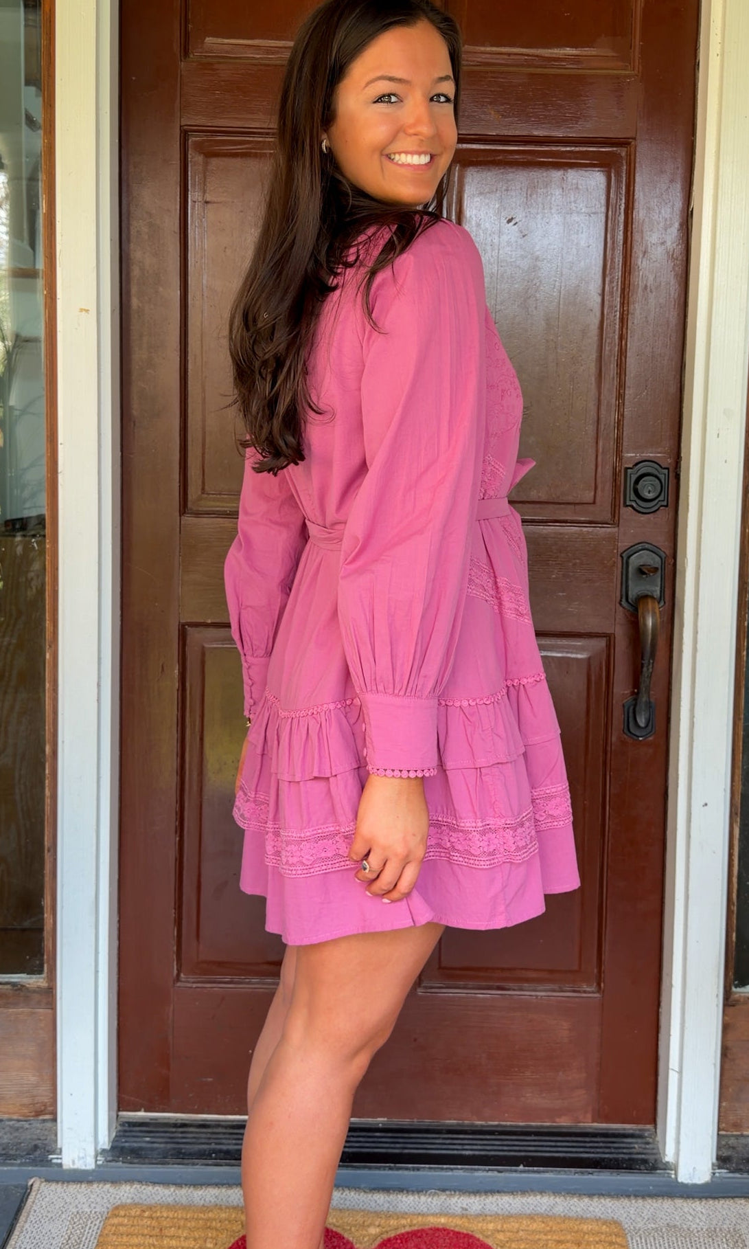 Mauve high-neck lace trim mini dress with long sleeves, self-tie waist, and tiered ruffle skirt worn by model standing in front of a wooden door