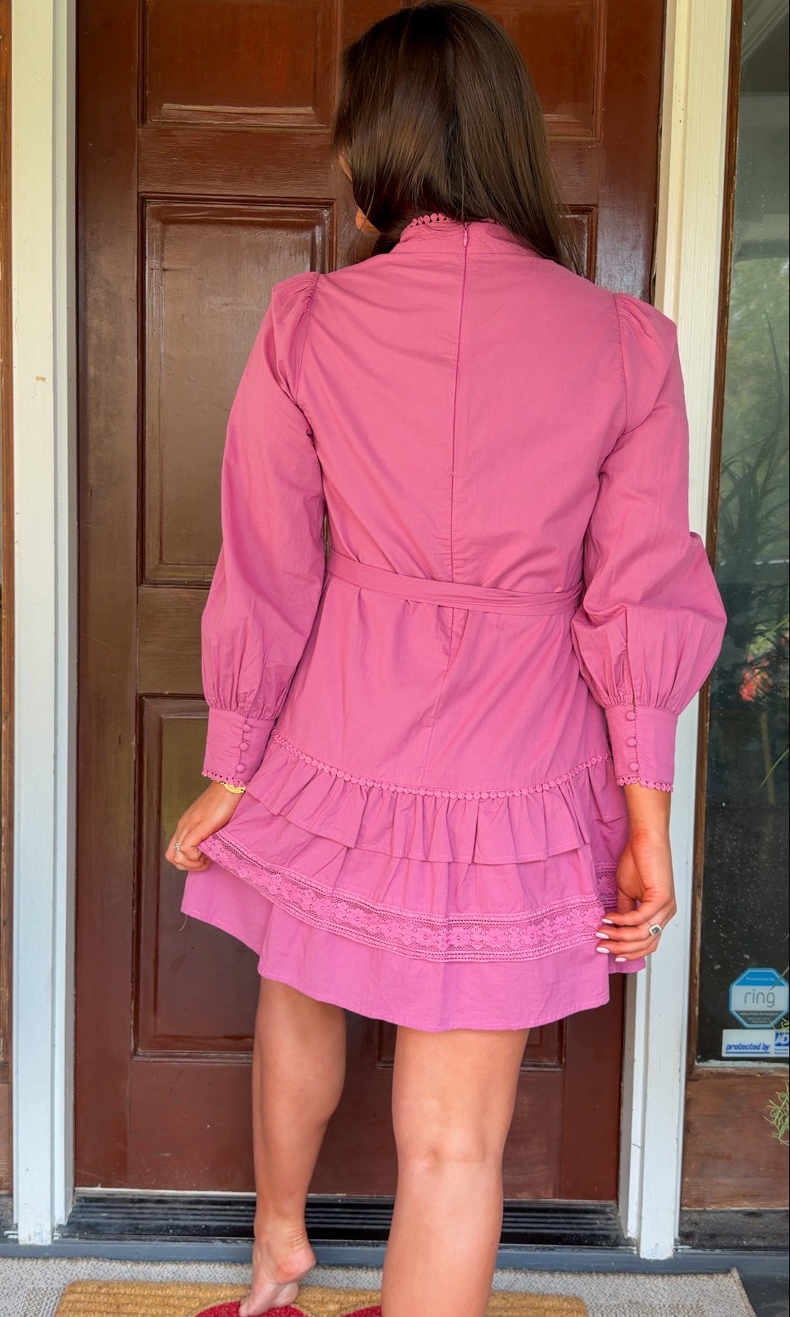 Mauve high-neck lace trim mini dress with long sleeves, self-tie waist, and tiered ruffle skirt worn by model standing in front of a wooden door