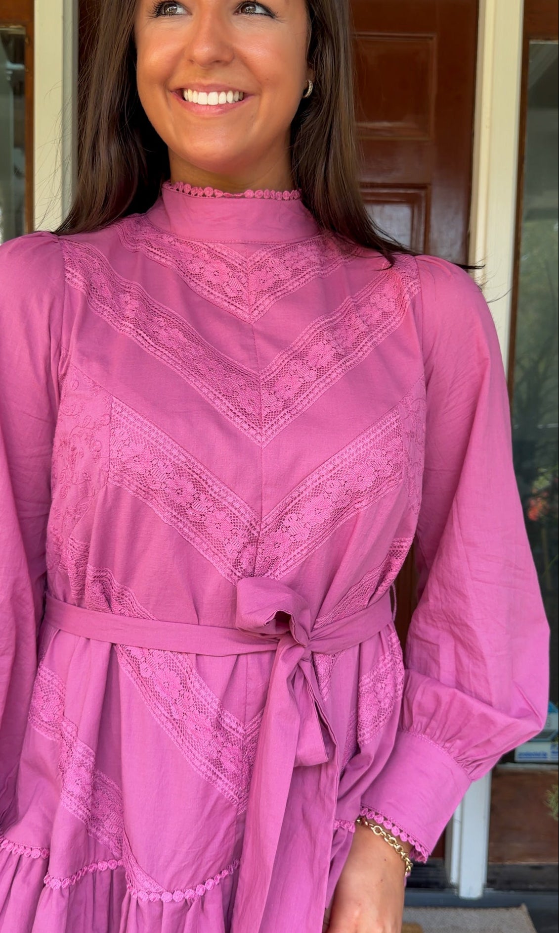 Mauve high-neck lace trim mini dress with long sleeves, self-tie waist, and tiered ruffle skirt worn by model standing in front of a wooden door