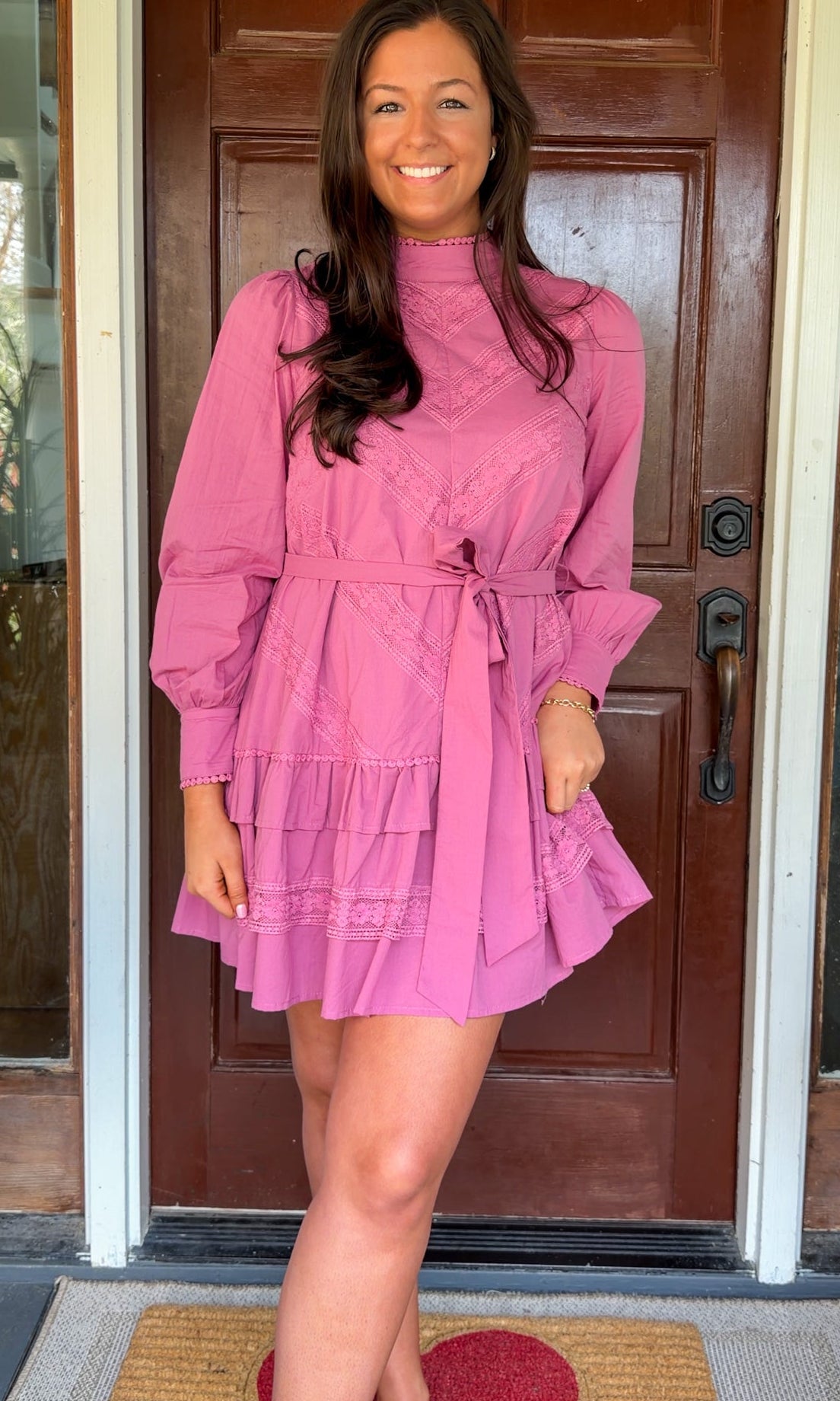 Mauve high-neck lace trim mini dress with long sleeves, self-tie waist, and tiered ruffle skirt worn by model standing in front of a wooden door