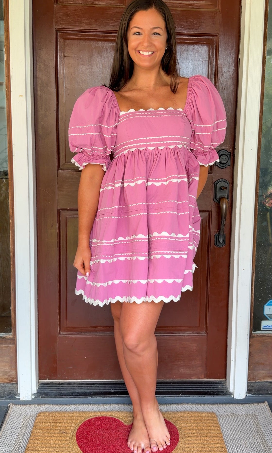 Model wearing a pink cotton poplin mini dress with white trim inset details, puff sleeves, and a square neckline, standing in front of a wooden door.