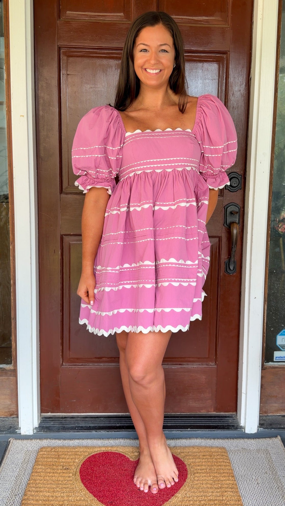 Model wearing a pink cotton poplin mini dress with white trim inset details, puff sleeves, and a square neckline, standing in front of a wooden door.