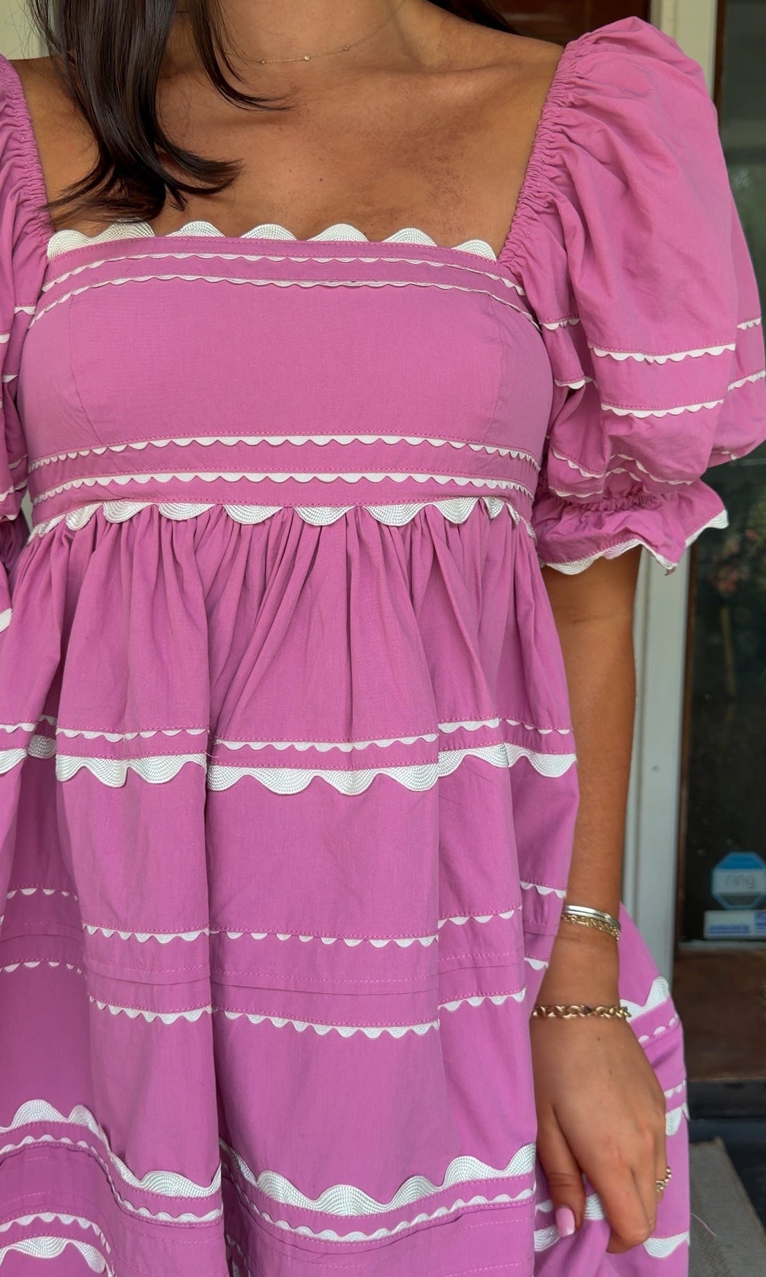 Model wearing a pink cotton poplin mini dress with white trim inset details, puff sleeves, and a square neckline, standing in front of a wooden door.
