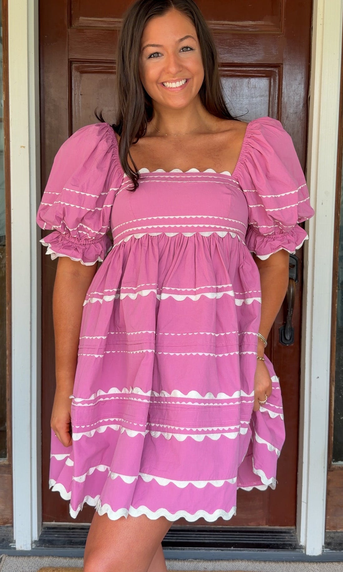 Model wearing a pink cotton poplin mini dress with white trim inset details, puff sleeves, and a square neckline, standing in front of a wooden door.