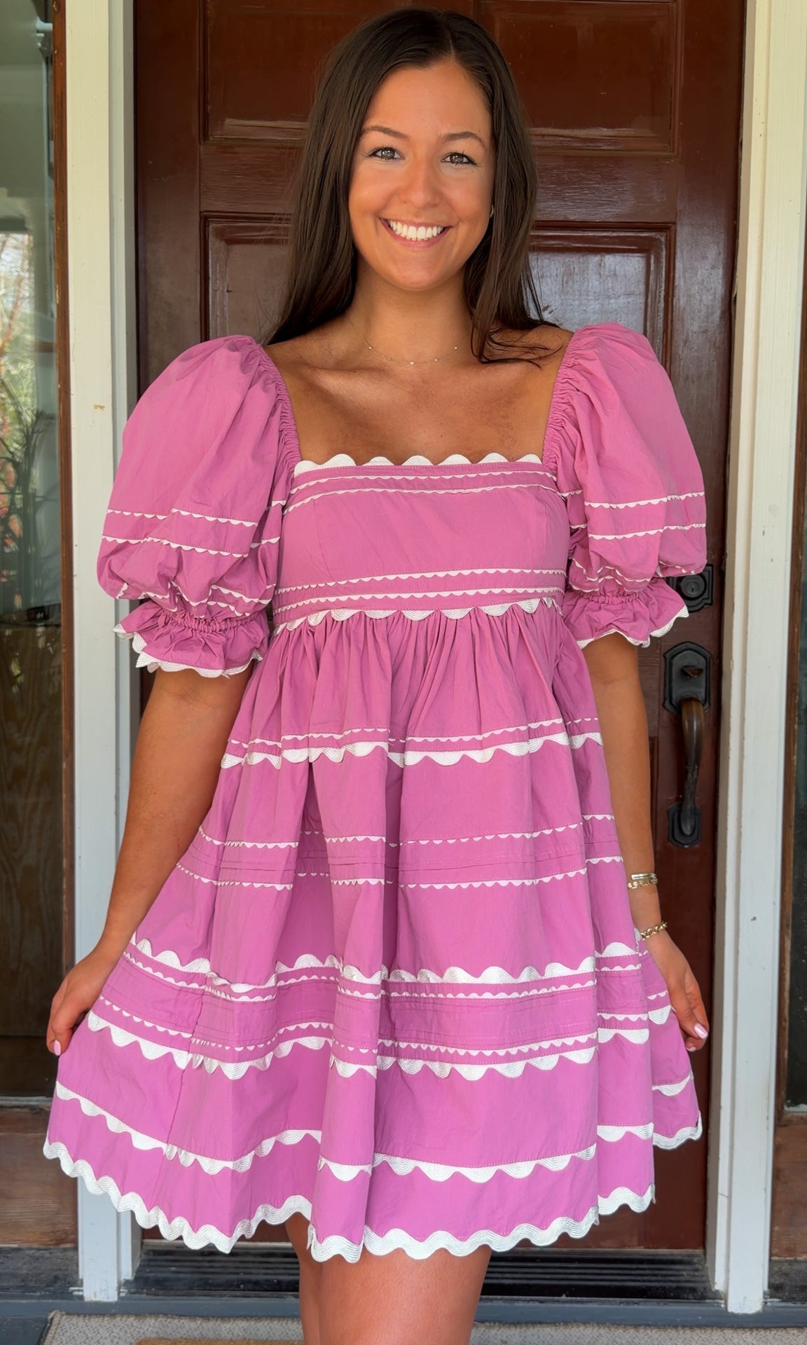 Model wearing a pink cotton poplin mini dress with white trim inset details, puff sleeves, and a square neckline, standing in front of a wooden door.