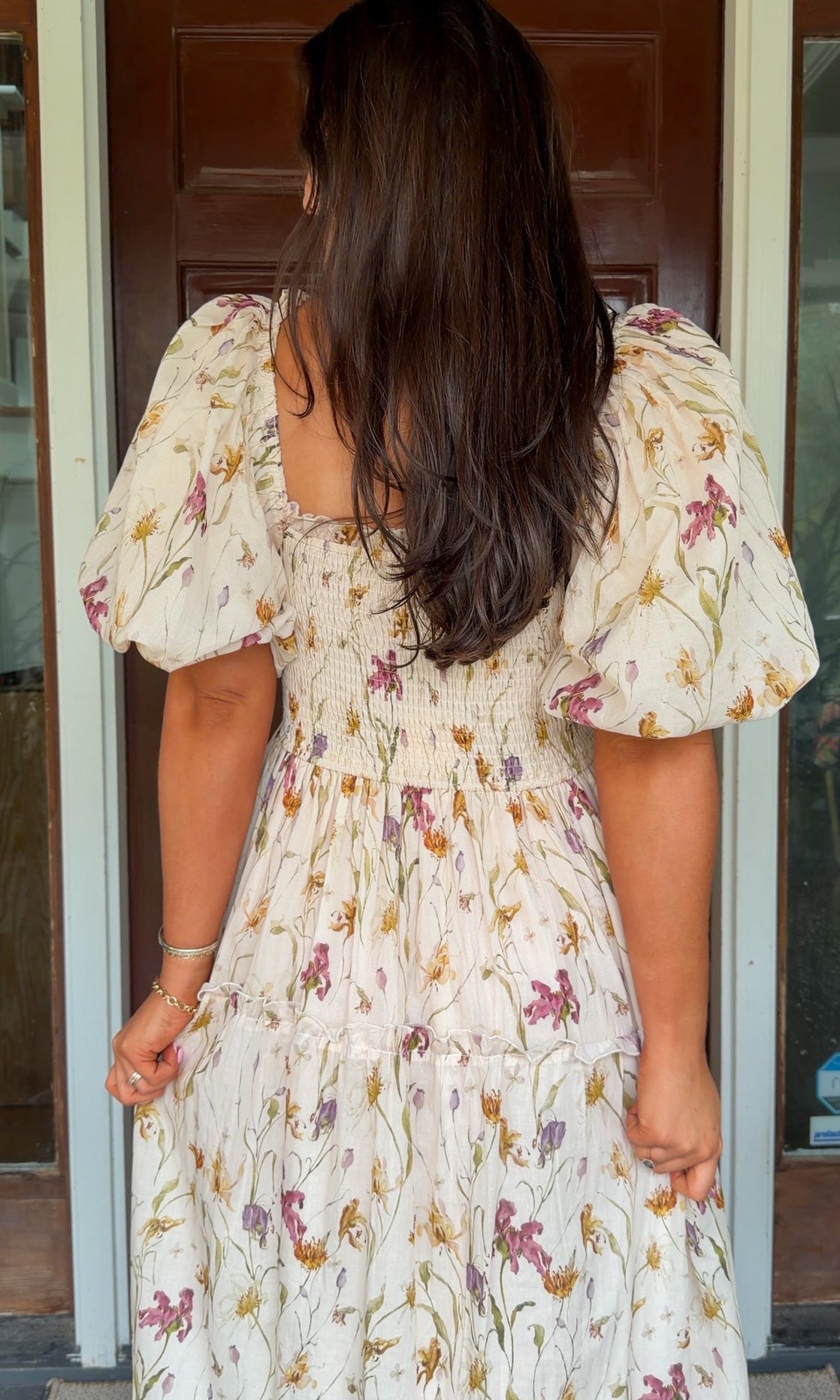 Model wearing a cream floral smocked midi dress with puff sleeves, flowy skirt, and side pockets, photographed in front of a wooden door.