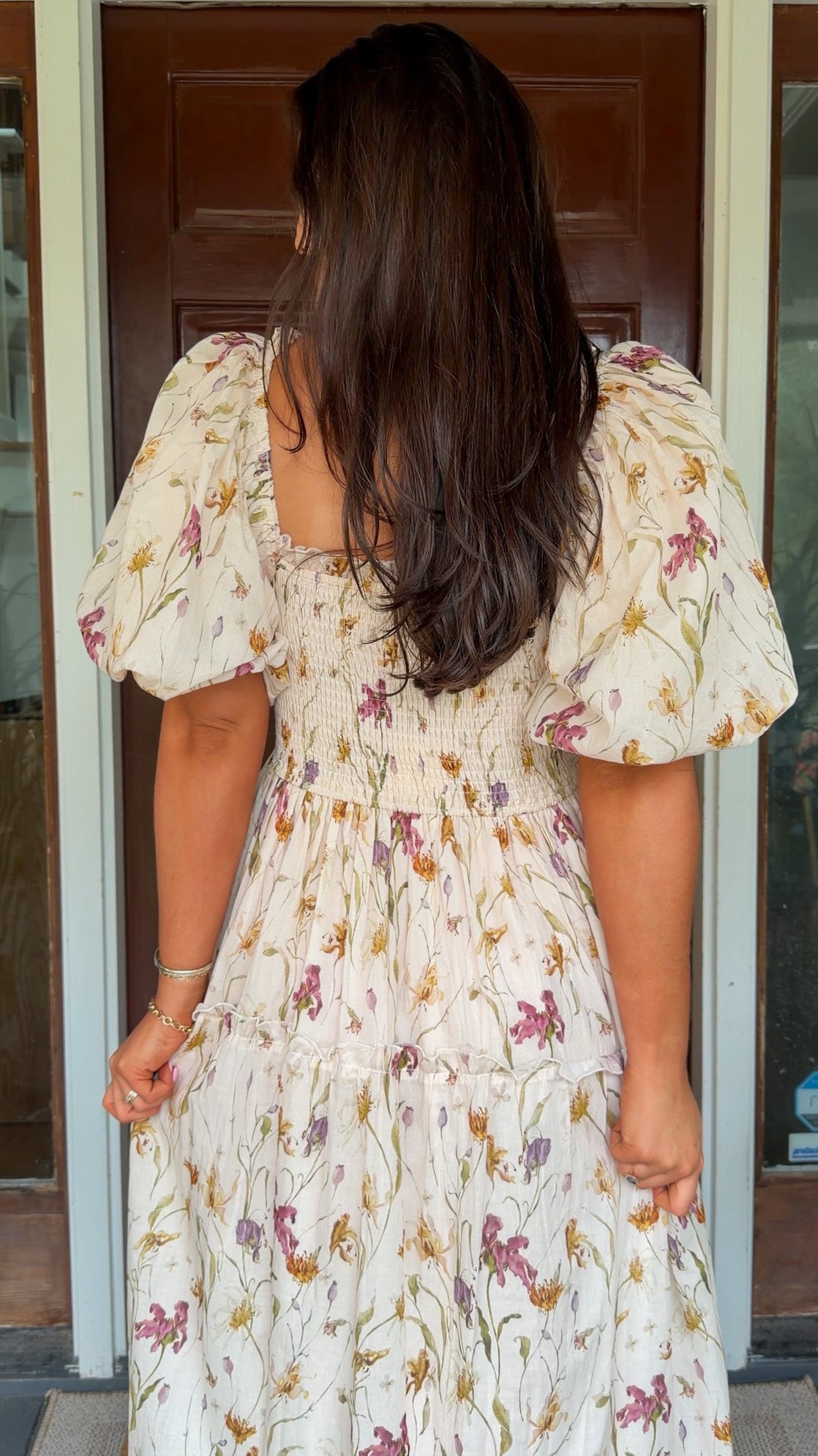 Model wearing a cream floral smocked midi dress with puff sleeves, flowy skirt, and side pockets, photographed in front of a wooden door.