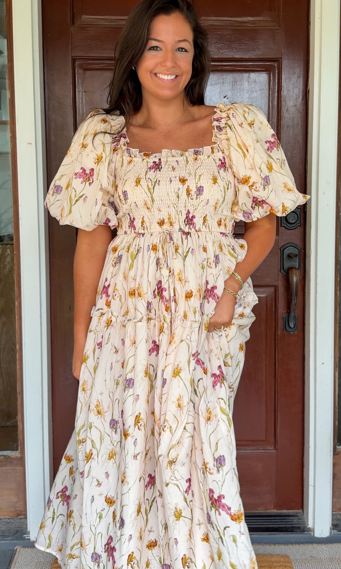 Model wearing a cream floral smocked midi dress with puff sleeves, flowy skirt, and side pockets, photographed in front of a wooden door.