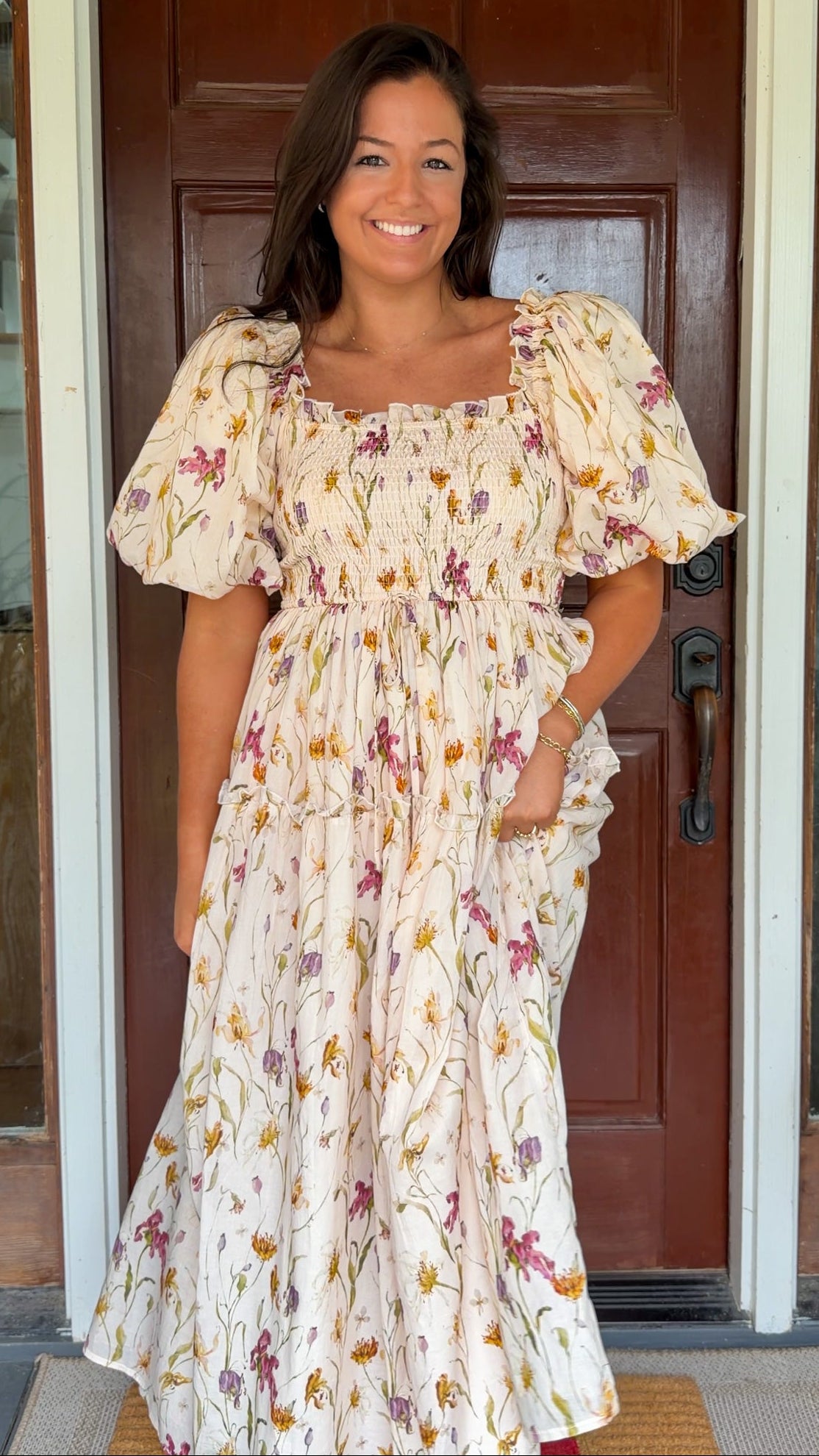 Model wearing a cream floral smocked midi dress with puff sleeves, flowy skirt, and side pockets, photographed in front of a wooden door.
