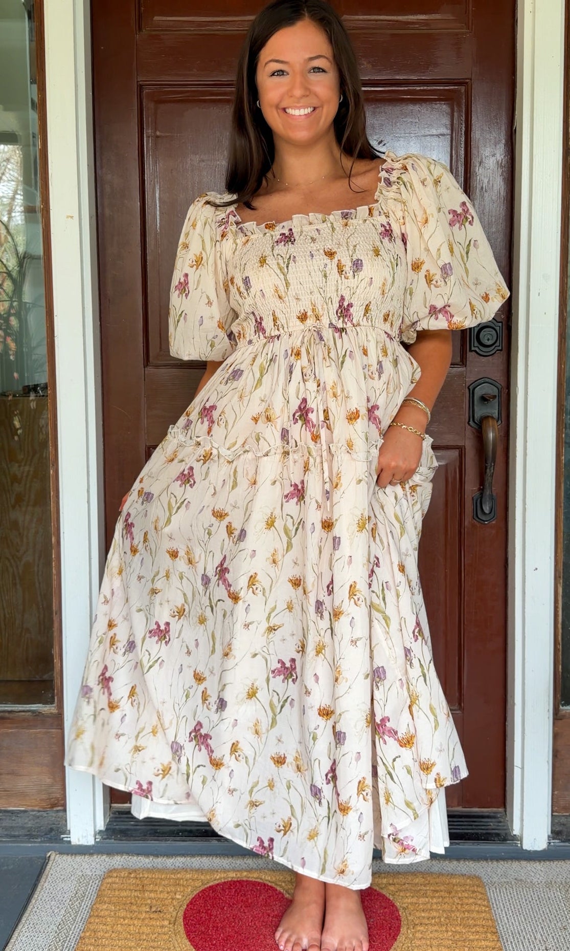 Model wearing a cream floral smocked midi dress with puff sleeves, flowy skirt, and side pockets, photographed in front of a wooden door.
