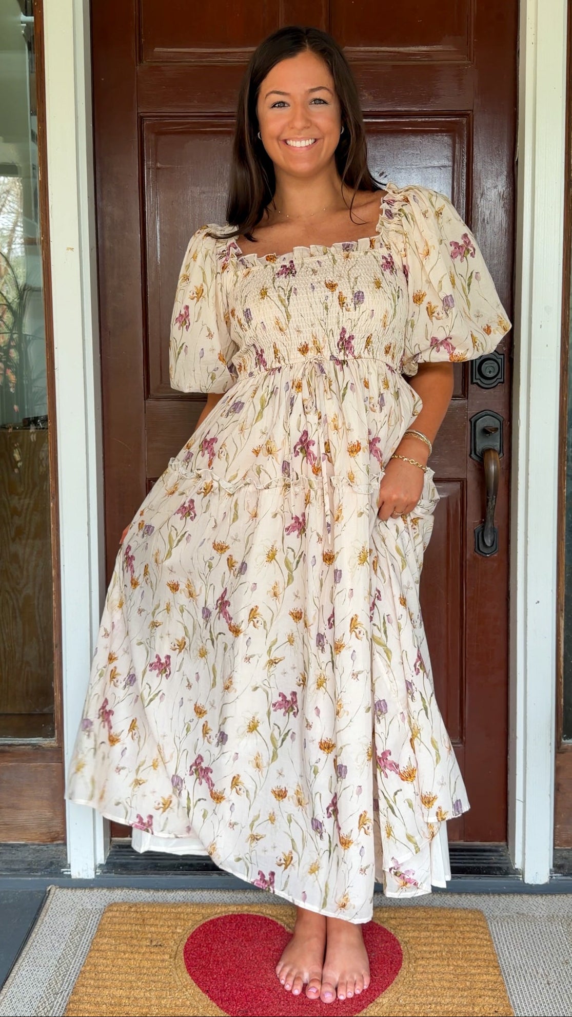 Model wearing a cream floral smocked midi dress with puff sleeves, flowy skirt, and side pockets, photographed in front of a wooden door.