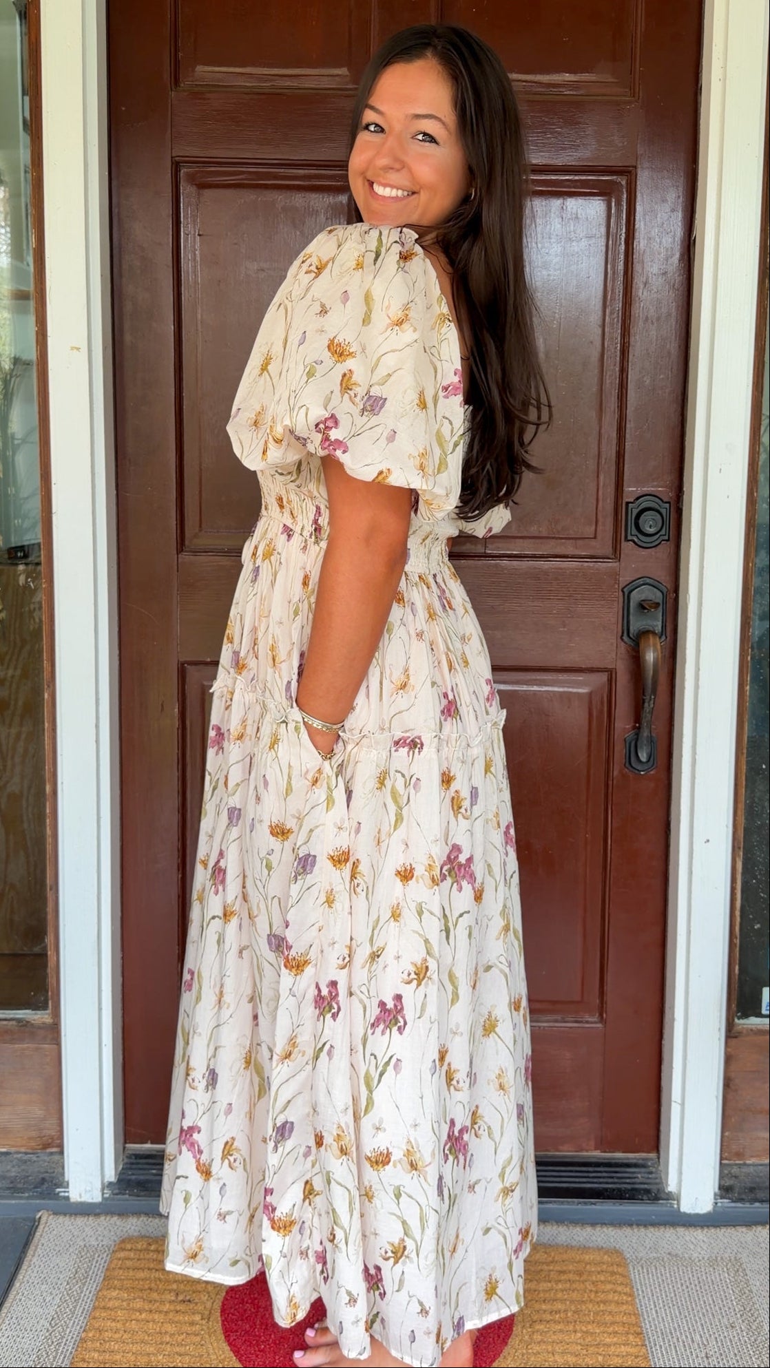 Model wearing a cream floral smocked midi dress with puff sleeves, flowy skirt, and side pockets, photographed in front of a wooden door.