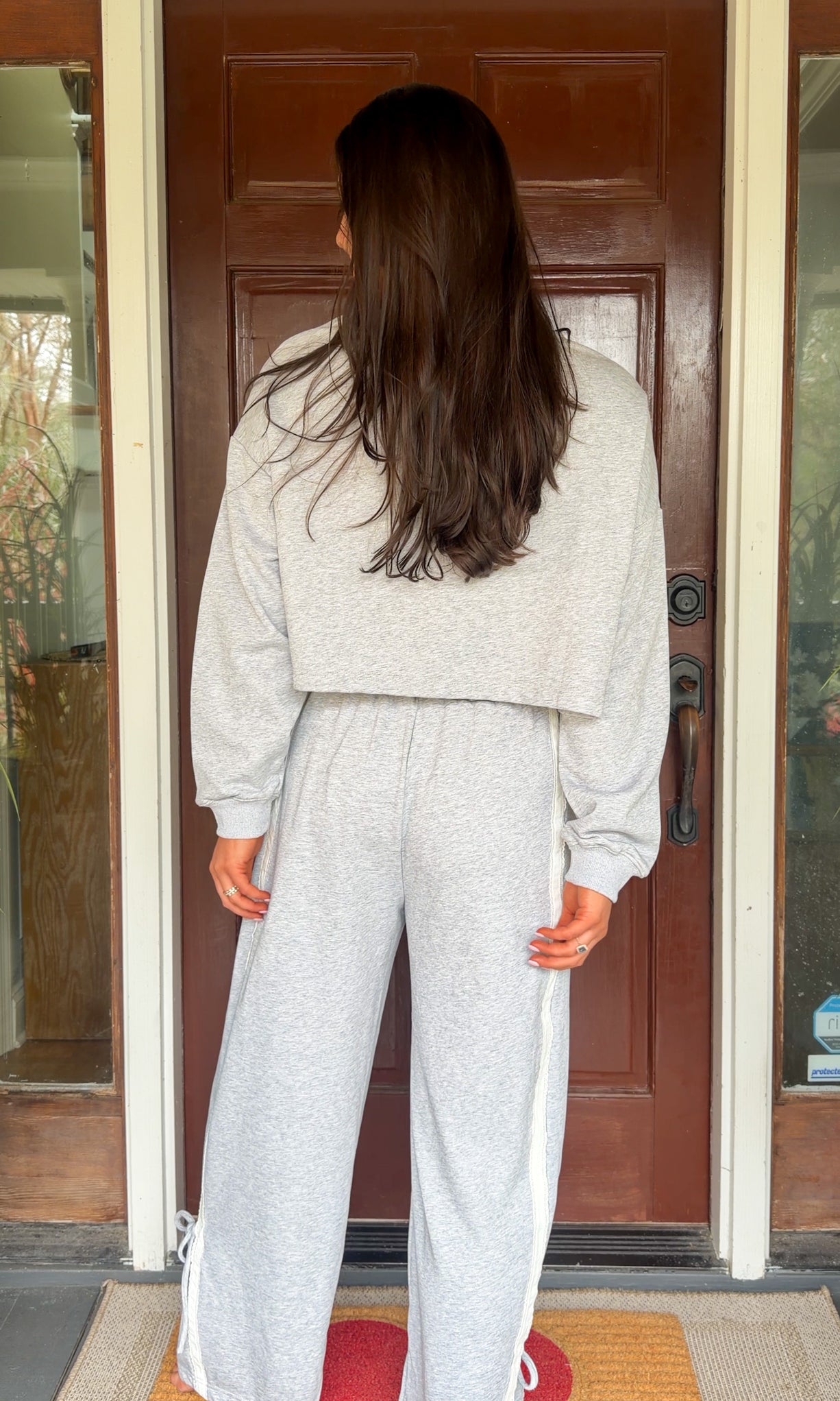 Model wearing a gray two-piece lounge set with lace trim cropped sweatshirt and matching elastic waist track pants featuring white side stripes and bow tie ankle details, standing in front of a wooden door.