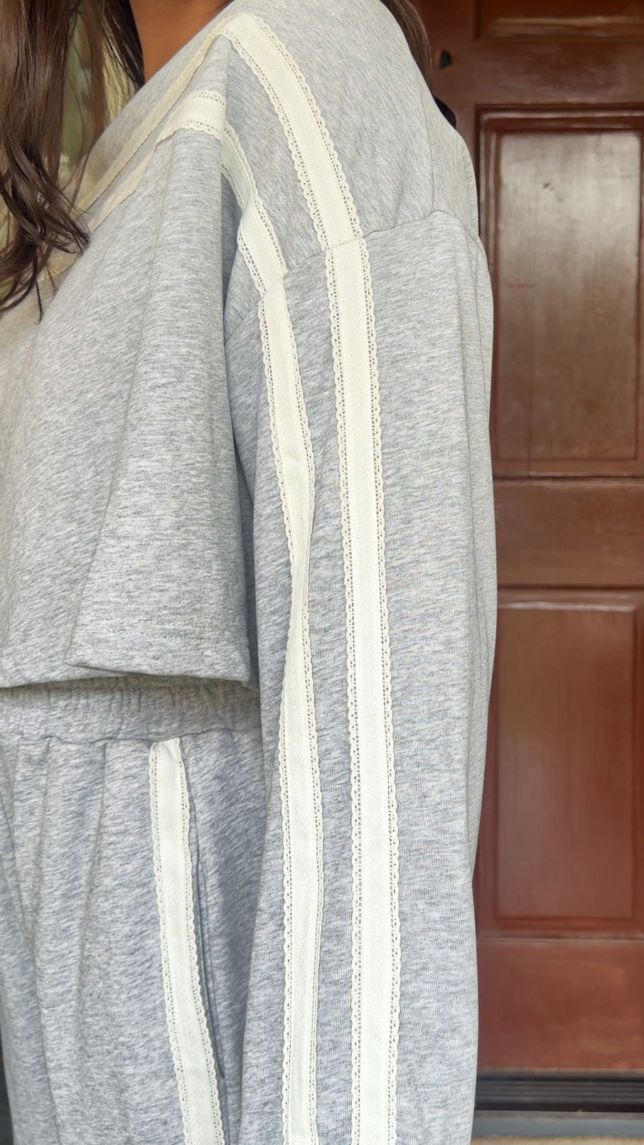 Model wearing a gray two-piece lounge set with lace trim cropped sweatshirt and matching elastic waist track pants featuring white side stripes and bow tie ankle details, standing in front of a wooden door.