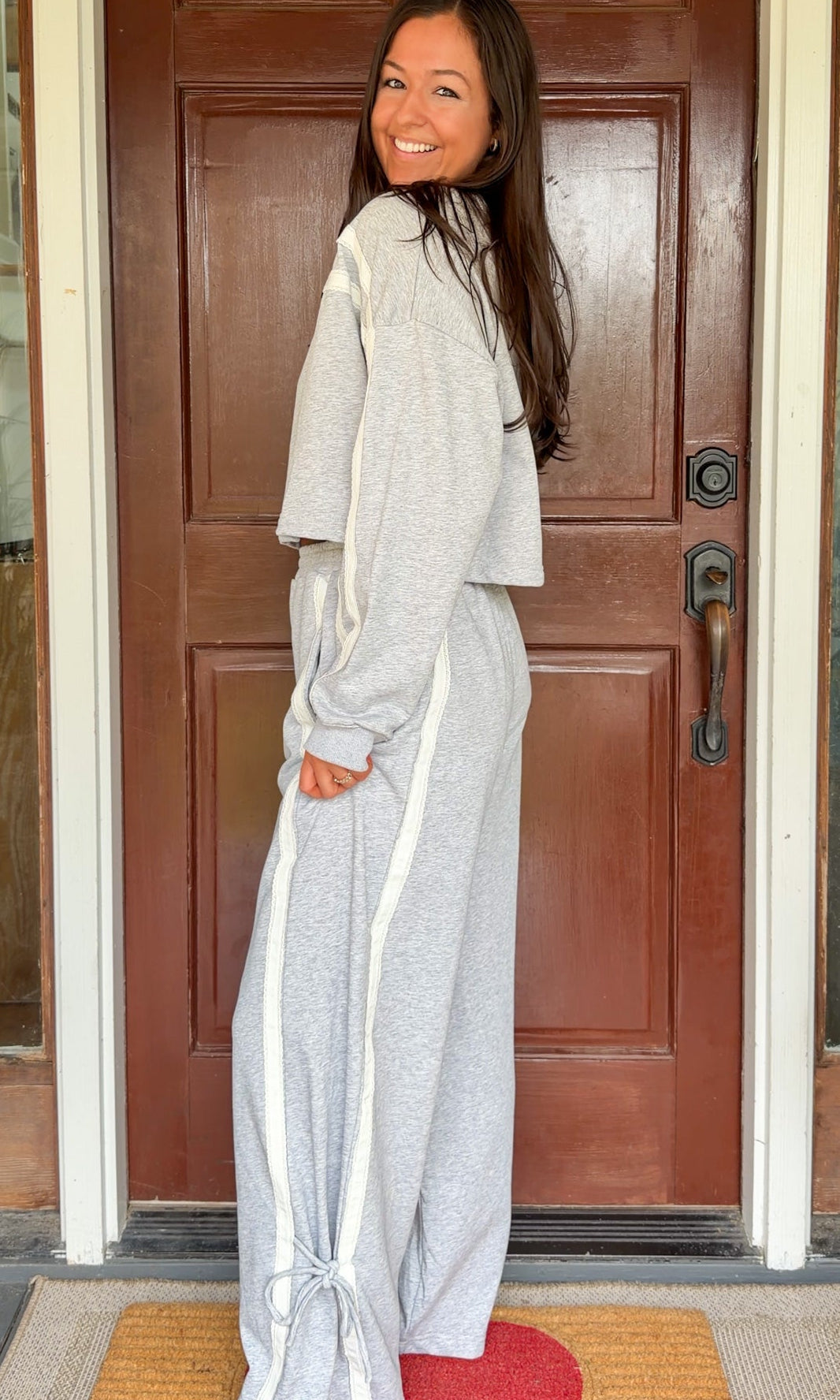 Model wearing a gray two-piece lounge set with lace trim cropped sweatshirt and matching elastic waist track pants featuring white side stripes and bow tie ankle details, standing in front of a wooden door.