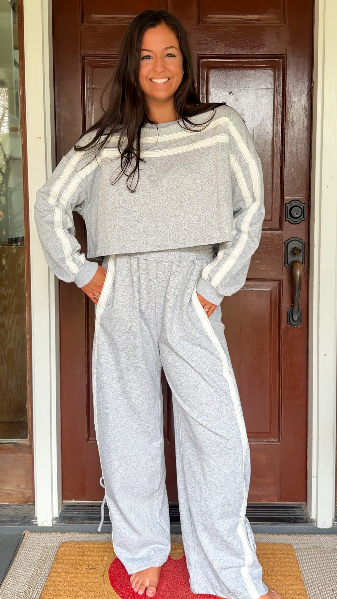 Model wearing a gray two-piece lounge set with lace trim cropped sweatshirt and matching elastic waist track pants featuring white side stripes and bow tie ankle details, standing in front of a wooden door.