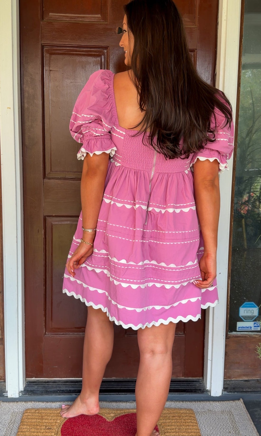 Model wearing a pink cotton poplin mini dress with white trim inset details, puff sleeves, and a square neckline, standing in front of a wooden door.